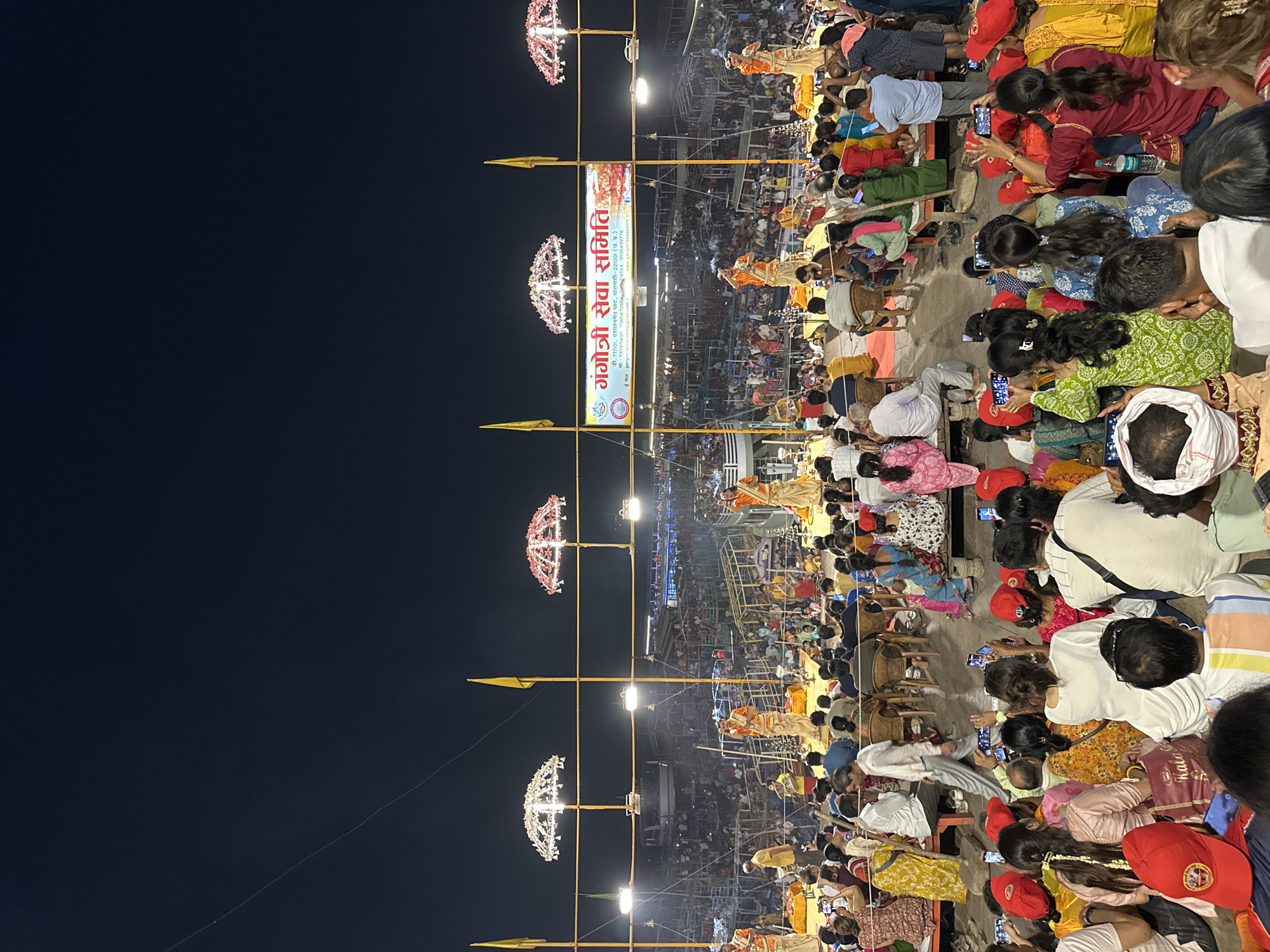 The image captures a densely packed crowd gathered in Varanasi, India, likely at the Dashashwamedh Ghat, a prominent riverfront location. The evening sky is dark. The focus is on a religious ceremony or gathering, where figures in orange robes can be seen on raised platforms, suggesting they are priests or performers. In the foreground, many attendees are seated on the ground, most of them holding up their phones to record or photograph the event. Vertical poles decorated with illuminated ornaments and flags are spaced out, leading the eye toward a banner with the words "गंगोत्री सेवा समिति" in Hindi, implying a religious or community organization is involved. A metallic structure in the background, possibly a building, is partly visible amidst the crowd. The scene illustrates a public religious event, with the attendees actively documenting the proceedings with their mobile devices.