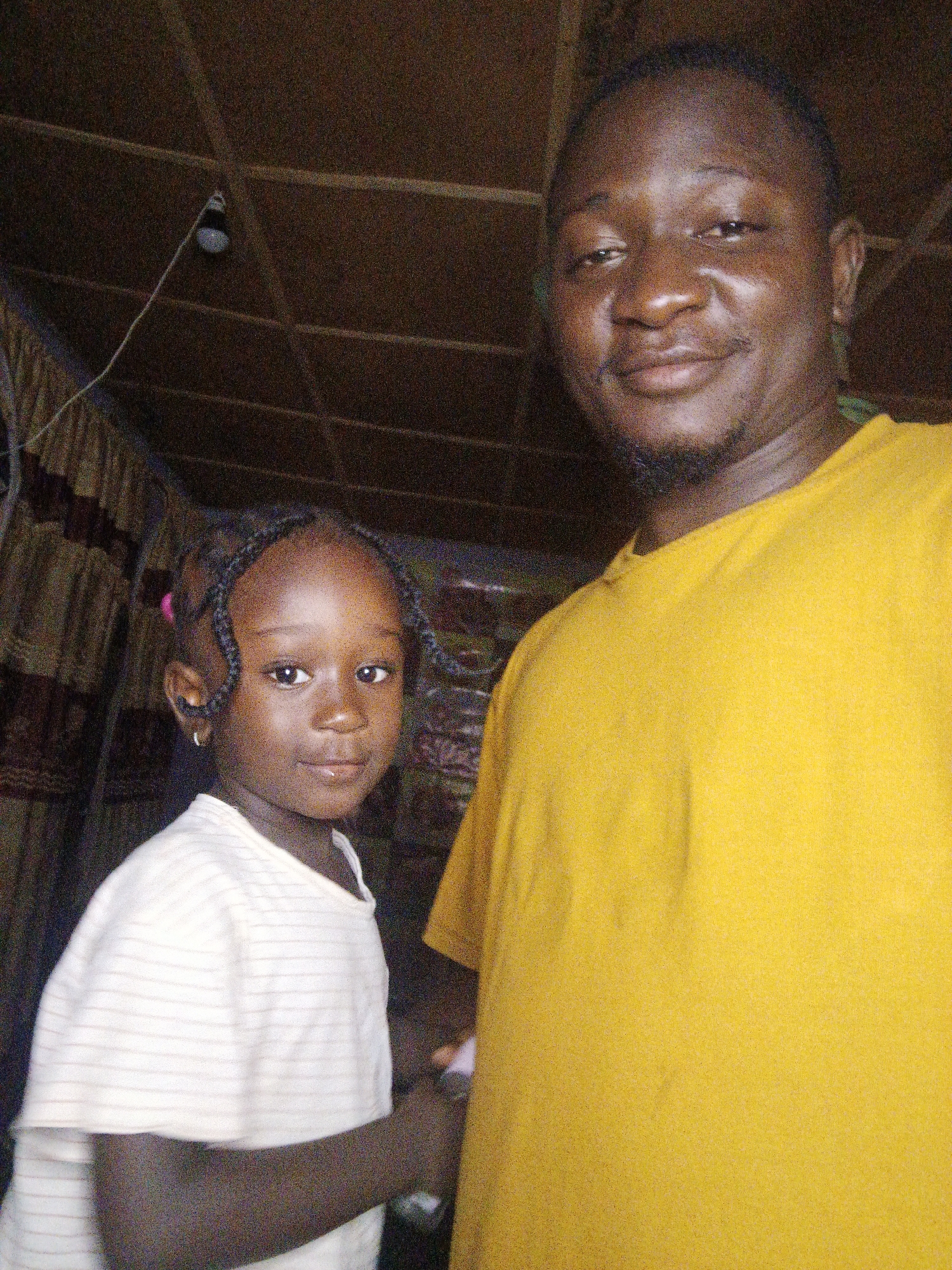 The image captures an outdoor scene, possibly in a rural or informal urban setting. The background reveals a rustic ceiling made of wooden beams and planks, with a single light bulb suspended from a wire. To the left, a patterned curtain or fabric hangs, suggesting a makeshift dwelling or shelter.
In the foreground, a man and a young girl are posing for the photo. The man, on the right, is wearing a bright yellow t-shirt and has a warm expression. The young girl, on the left, is wearing a white striped t-shirt and has her hair styled in braids. She is looking directly at the camera with a serious expression. The overall lighting suggests it is daytime, but the interior setting is somewhat dimly lit, possibly due to the overhead canopy. The image has a grainy quality, indicative of a lower resolution or older camera. The interaction between the two subjects appears to be a posed portrait, likely taken by a third party.