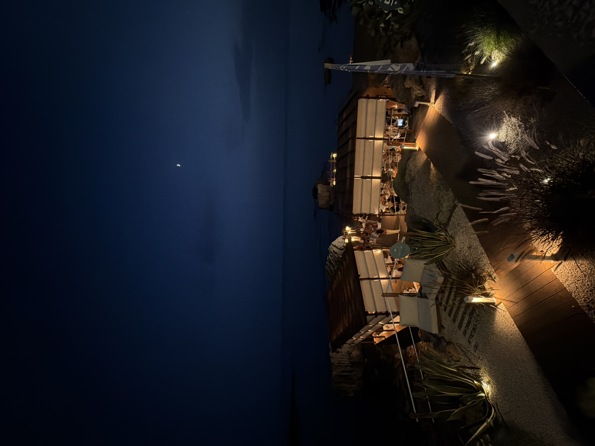 This is a nighttime, high-angle, outdoor, wide shot of an illuminated open-air restaurant situated on a rocky coastline near the city of Alghero, Italy. The dark blue sky is visible above, with a sliver of a crescent moon and a small bright star. The sea stretches out to the horizon, appearing dark and calm.
The restaurant features two connected, rectangular structures with white canvas roofs supported by wooden beams. The structures are illuminated from within by warm, soft lighting, casting a glow on the tables and chairs where approximately 15-20 patrons are seated, appearing to be dining. The structures are set on a rocky elevation.
In the foreground, a wooden walkway, lit by strategically placed small, warm lights, curves through a landscape of light-colored gravel and sparse vegetation. Large-leafed plants, possibly agave, are visible to the left, illuminated by ground lights. Tall, feathery grasses are present in the mid-ground and right, also lit from below. Two white lounge chairs are placed on the gravel area near the wooden walkway. A white flag with blue markings is affixed to a pole on the right side of the image. The overall atmosphere is serene and inviting, suggesting a tranquil dining experience by the sea.
