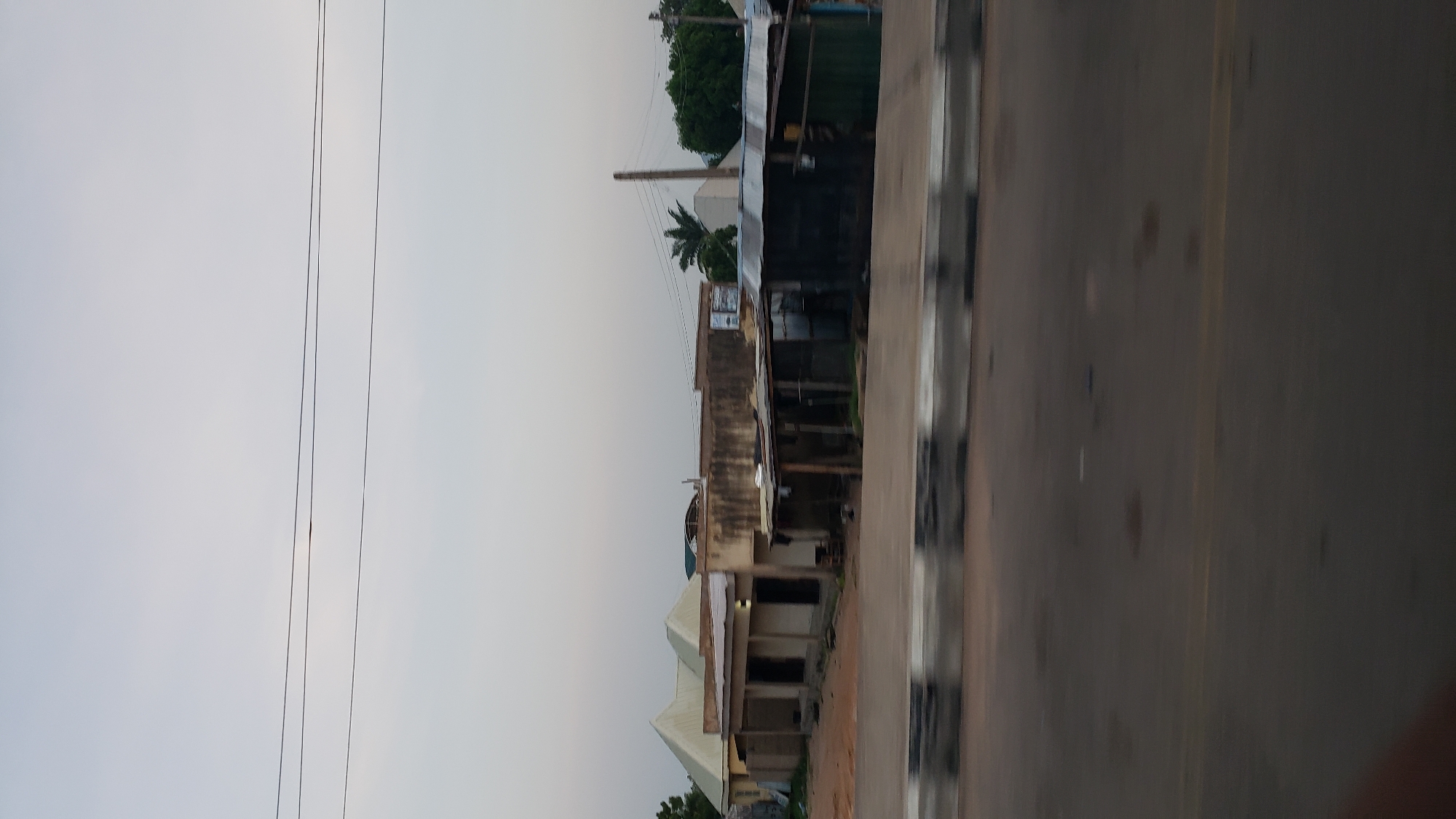 The image captures a street scene in Jalingo, Nigeria, on what appears to be a slightly overcast day. In the mid-ground, a row of buildings is visible. To the left, a structure with a distinctive double-pitched roof is under construction or renovation, its walls unpainted. Adjacent to it, a longer, single-story building with multiple entrances, possibly shops, has an open-air, covered walkway in front. Further to the right, a series of smaller, informal structures with corrugated metal roofs and open fronts are situated, appearing somewhat makeshift. These structures seem to be commercial stalls or small businesses. A single utility pole with visible wires stands behind these structures. In the foreground, a road with a distinct yellow line and a concrete barrier with black and white markings runs horizontally across the frame, indicating a street or highway. The background consists of a pale sky and trees on the right side, suggesting a suburban or semi-urban setting within Jalingo. There are no people clearly visible in the image, and the scene is static.