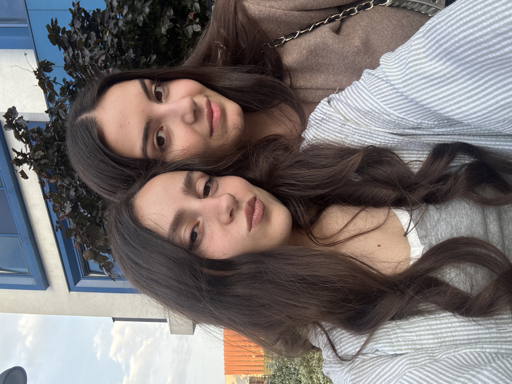 The image is a selfie-style portrait of two young women, taken outdoors and rotated 90 degrees counter-clockwise.
The woman in the foreground has long, wavy dark brown hair, fair skin, and looks towards the camera with a neutral, slightly pleasant expression. A small mole is visible on her upper chest. She is wearing a light-colored top with thin horizontal stripes in light green/grey and white. Her hair flows around her shoulders and chest.
Behind her, the second woman's face is partially visible, showing her dark eyes and nose, also looking towards the camera with a neutral expression. She likewise has long, dark brown hair and is wearing a brown, ribbed, long-sleeved garment, possibly a sweater or jacket. A black and gold chain strap, likely from a bag, rests on her shoulder.
The background shows an outdoor setting with architectural and natural elements. To the left, a modern building with white walls and distinct blue window frames is visible, partially obscured by a dense bush or tree with dark leaves. Above, a blue sky with scattered white clouds is present. In the lower right background, an orange-tiled roof and distant structures or greenery can be faintly seen, suggesting an urban or suburban environment.
The women appear to be posing for a casual photograph, likely a selfie, on a clear daytime. No text is visible in the image. The location context provided is Opole, Poland.
