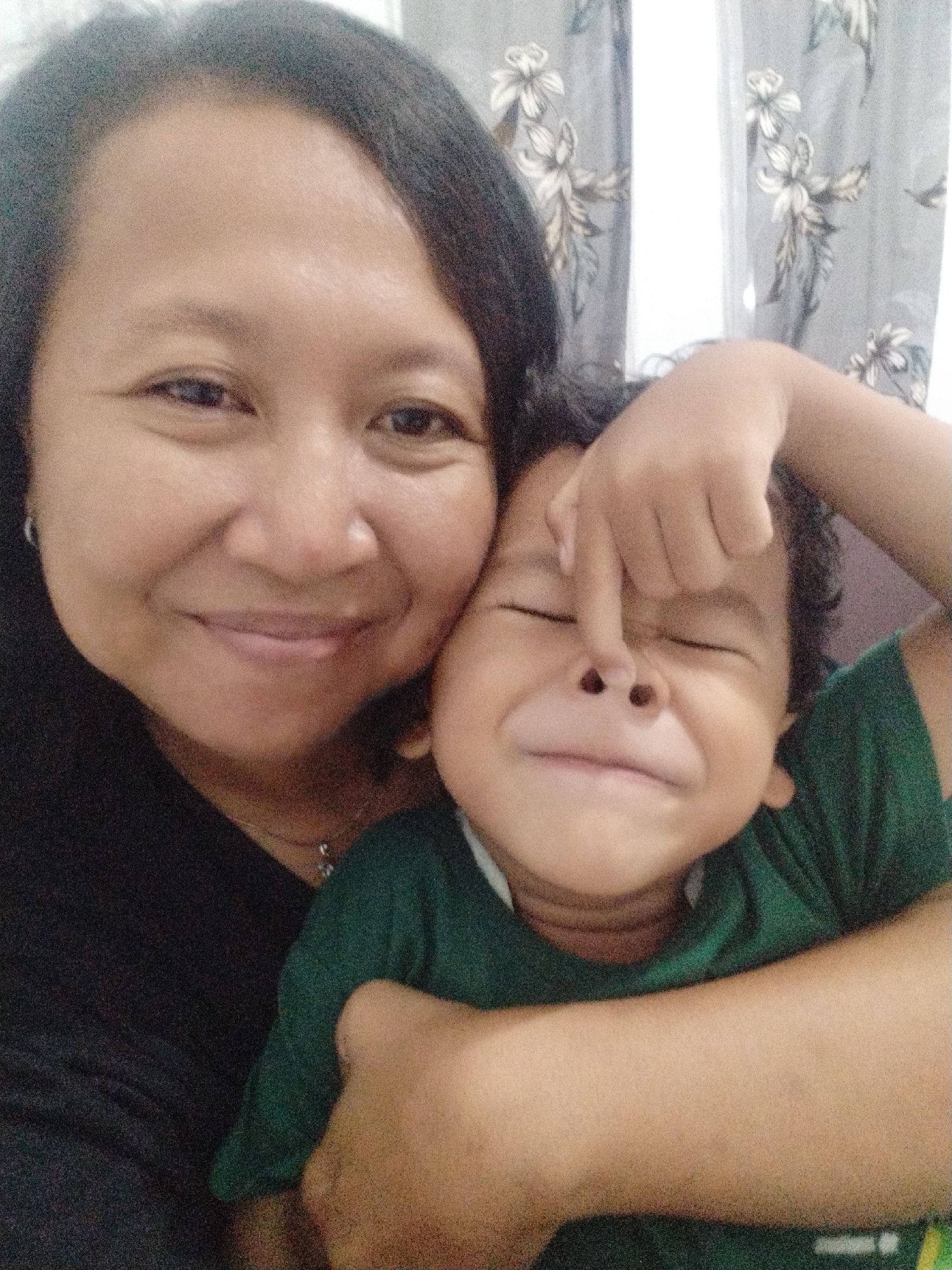 The image is a selfie of two individuals, an adult and a child, in what appears to be a domestic indoor setting in Karangsentosa, Indonesia. The adult, on the left, is a woman with dark hair, smiling at the camera. She is wearing a black top and a necklace. The child, positioned in front of the woman, is a young boy also facing the camera. He is wearing a green t-shirt, and his eyes are closed. His nose is being playfully touched by his own finger, and his mouth is open in a wide smile. The background consists of floral patterned curtains. The lighting is soft and diffused, suggesting it is daytime. The overall mood of the image is affectionate and playful.