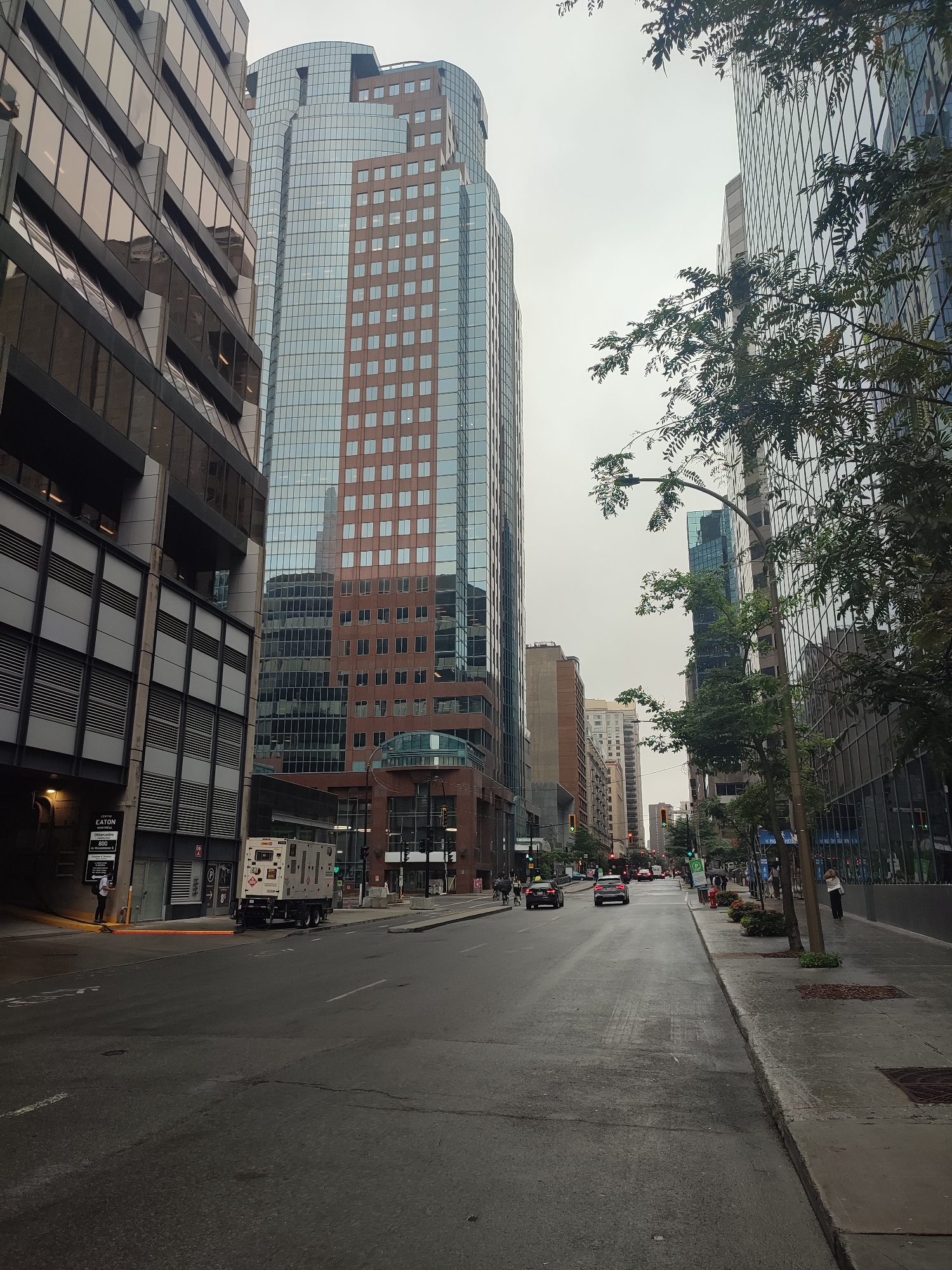 The image shows a street scene in Montreal, Canada, on a cloudy day. Tall, modern office buildings with glass facades dominate the left and right sides of the frame. The building on the left has a dark, textured exterior, while the central building is a mix of glass and reddish-brown brick. A white portable generator is parked on the side of the street in the foreground on the left. The street is wet, indicating recent rain or a damp day. Several cars are visible driving on the street, and a few pedestrians are walking on the sidewalk. Trees with green foliage are planted along the right side of the sidewalk. Signs for "CENTRE EATON MONTRÉAL" and parking are visible on the left. The overall atmosphere is urban and somewhat overcast.