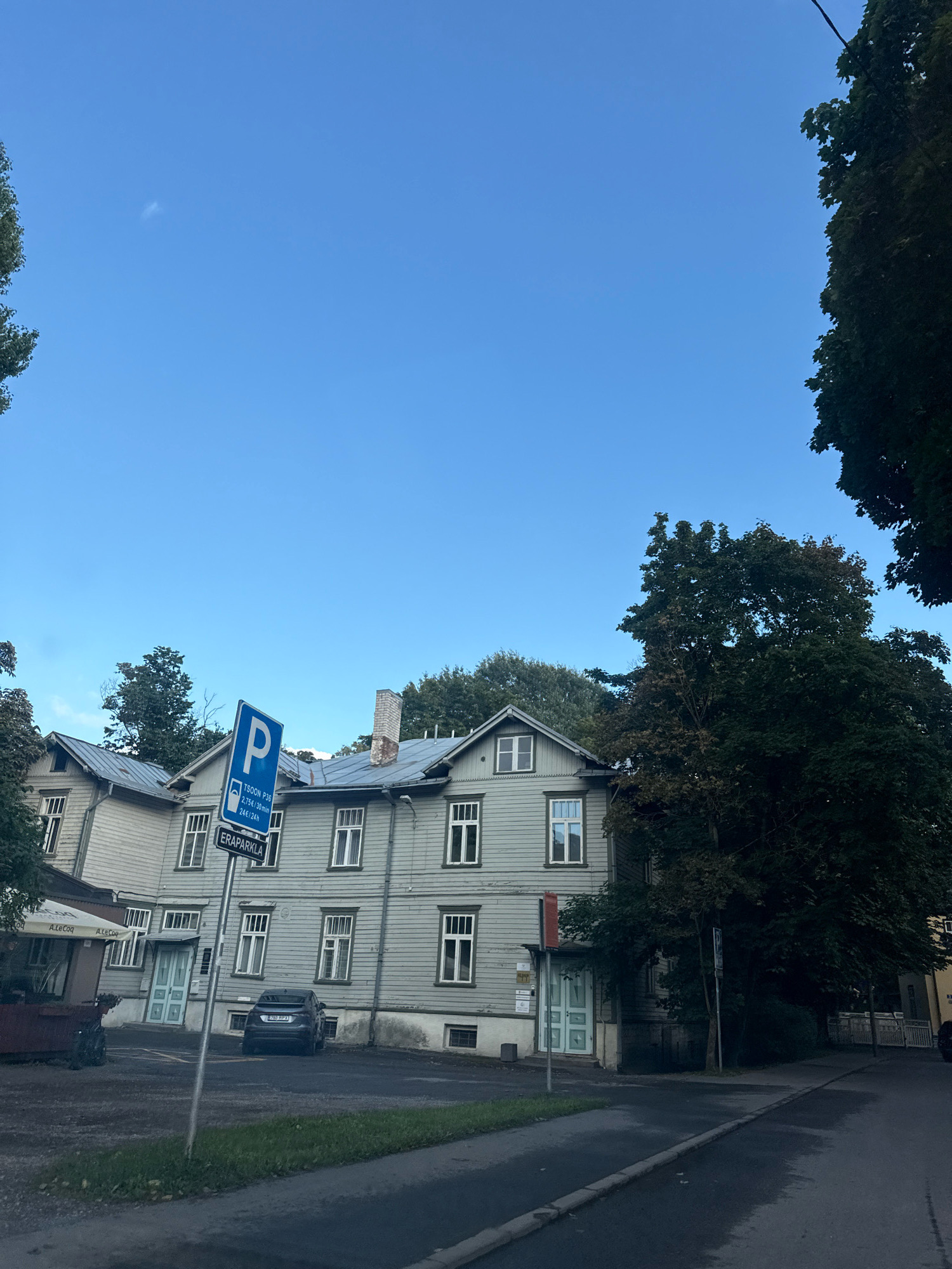 The image depicts a street scene in Tallinn, Estonia, with a focus on a pale green wooden building. The building appears to be two stories high with a pitched roof and several windows. A blue parking sign is prominently displayed in front of the building, indicating parking zone P36 with rates of 2.75 EUR per 30 minutes and 24 EUR per 24 hours, and the text "ERAPARKLA" which means private parking in Estonian. A dark-colored car is parked in front of the building. Trees surround the building, with a dense dark green tree on the right side of the image casting shadows. The sky is clear and blue. The foreground shows a paved road and a grassy area. The overall impression is an overcast day in a residential or mixed-use area of Tallinn.
