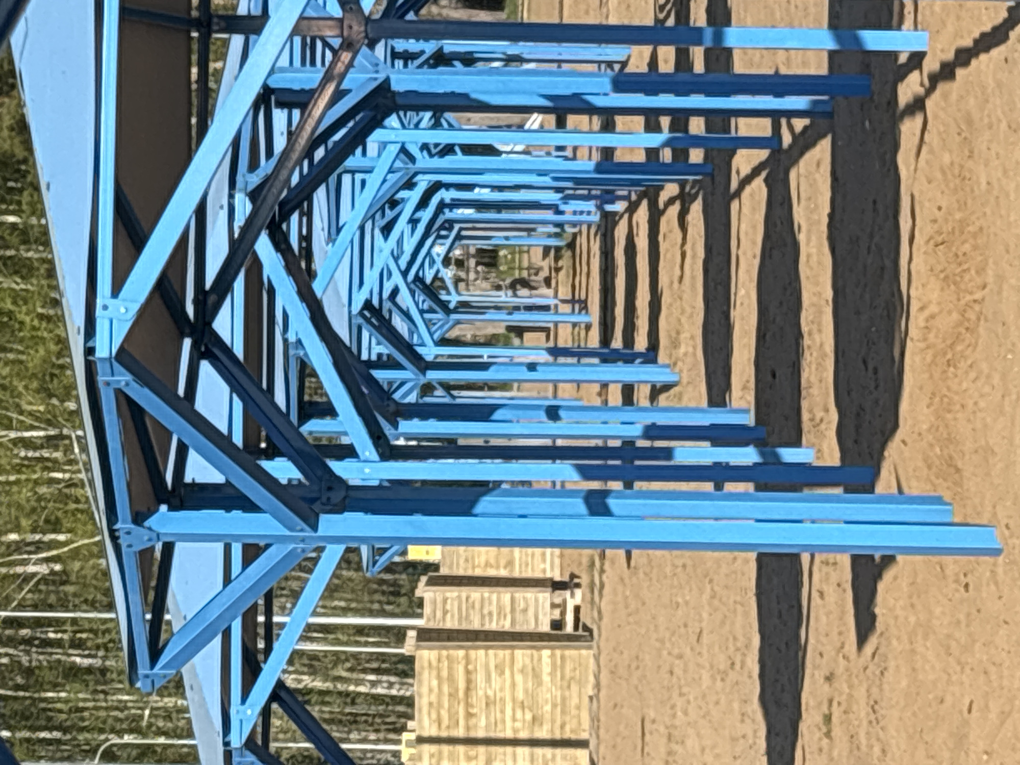A blue metal structure casts shadows on a tan surface. In the background, a wooded area and a light-colored building are visible. The location is Chaikovsky, Russia.