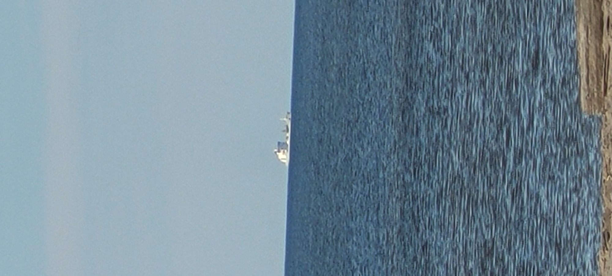 A distant cargo ship, predominantly white, is visible on the horizon over a dark blue body of water under a clear, pale blue sky. The water is slightly choppy, with small waves creating a textured surface. In the foreground, a light-colored sandy shore with a few pieces of wood is partially visible. The scene is calm and expansive, evoking a sense of open water and distance. The location is the coast of Riga, Latvia.