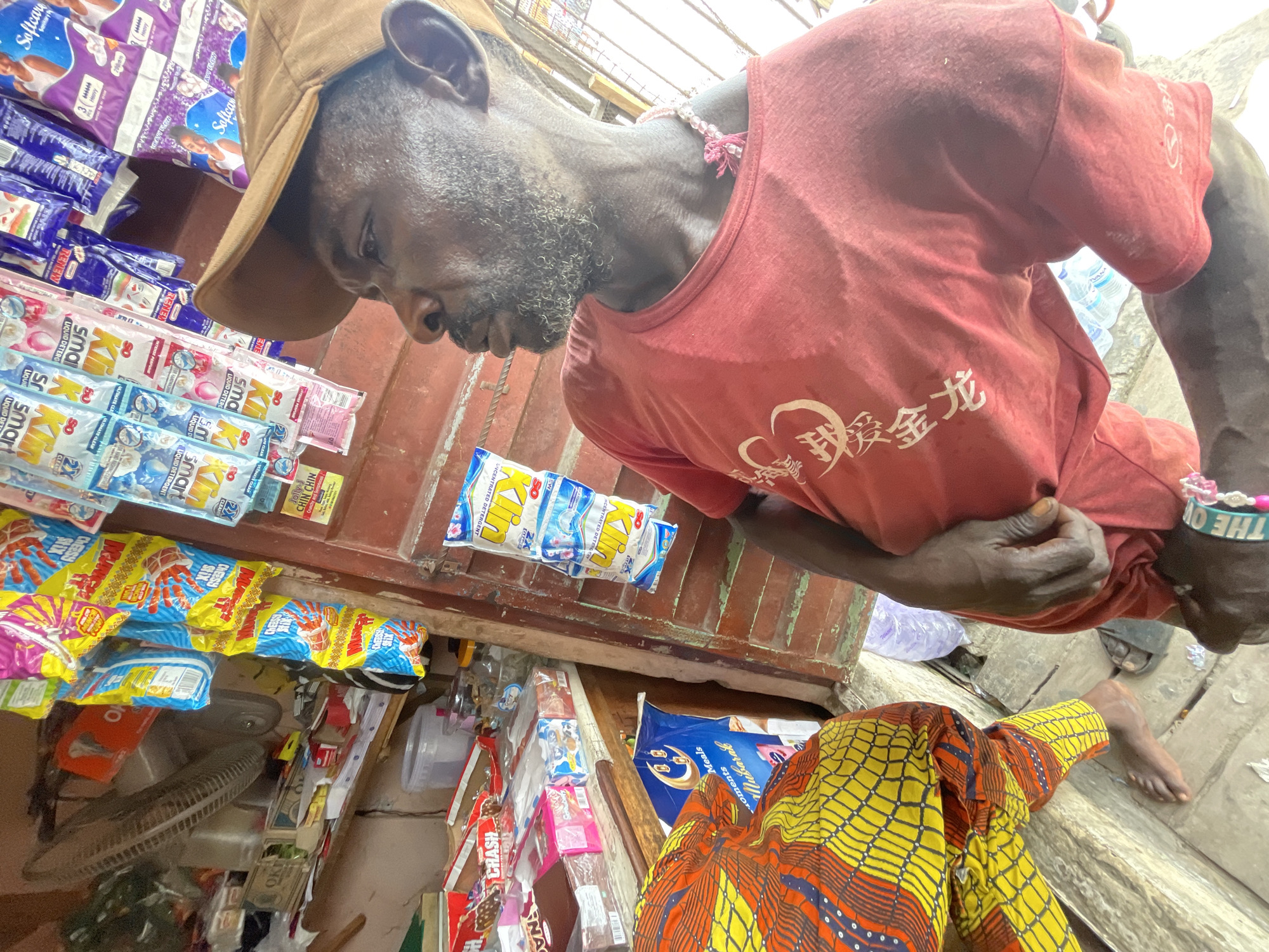 A street vendor in Ado, Nigeria, is captured in a high-angle medium shot. The man, wearing a brown cap and a red T-shirt with Chinese characters, is positioned behind a stall stocked with various household and food items. The stall showcases products like detergent (Softcare, King, Smart, Renew), snacks (Cheesy Stix, Jolly Chin Chin), and other grocery items. A small electric fan sits on the stall's lower shelf. The vendor is partially visible, with his bare feet and ankles exposed, as is a customer clad in a yellow and orange patterned fabric. The background suggests a bustling street scene with the presence of utility lines overhead.