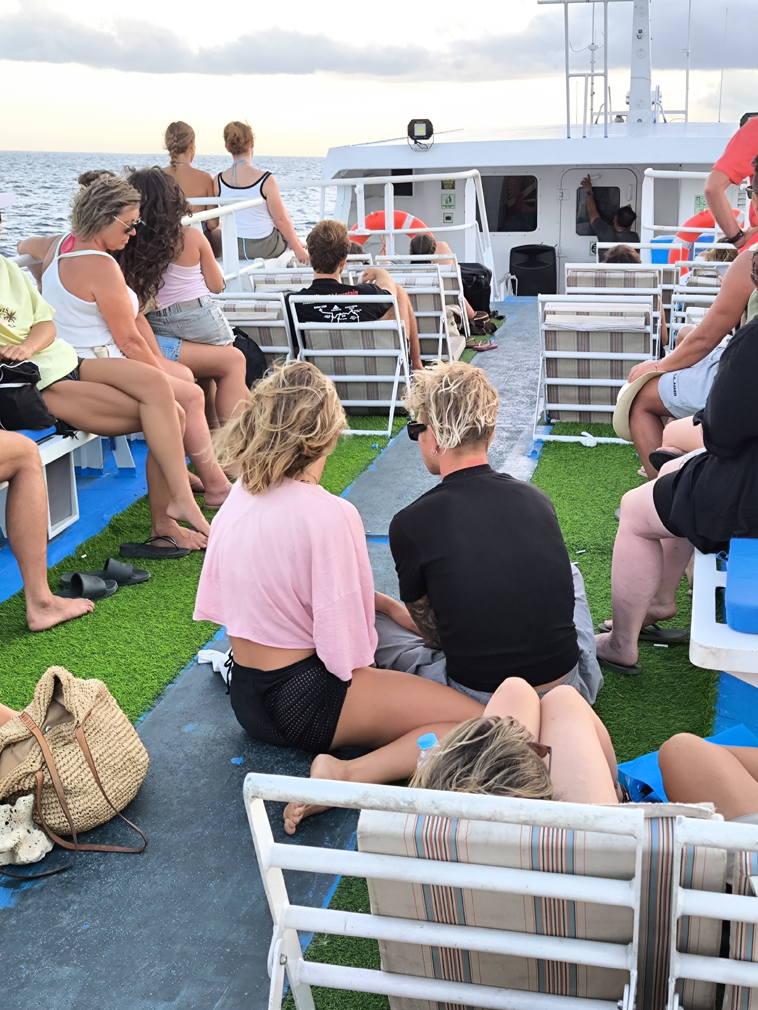 The image shows a group of people on a boat, likely a tour or excursion vessel. They are seated and standing on a deck covered in artificial turf. Most people are casually dressed in summer attire, such as shorts, tank tops, and t-shirts. Some are sitting in chairs while others are on the floor. There are life preservers visible on the boat's railing. The location is likely near the sea, given the horizon line in the background. The specific city and area in Indonesia is not identifiable from the image alone.