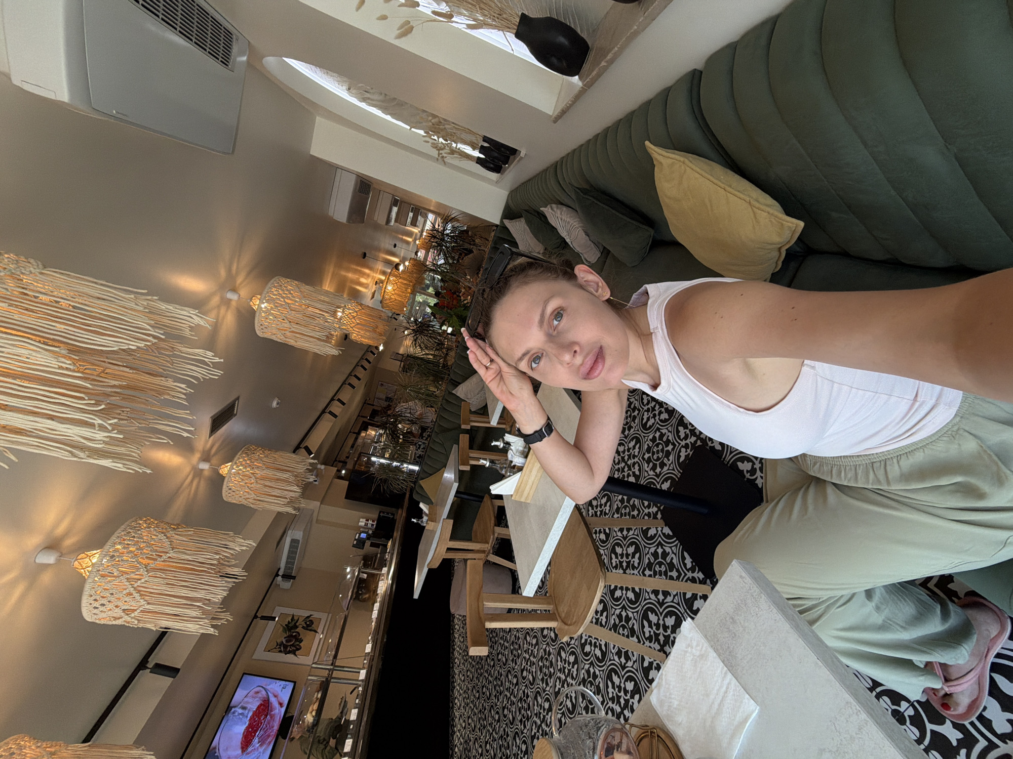 The image shows a woman taking a selfie in what appears to be a cafe or restaurant in Budva, Montenegro. The interior features a distinctive black and white patterned floor, wooden chairs with unique back designs, and a long, dark green tufted sofa. Above, macrame-style pendant lights with hanging fringe illuminate the space. A television screen displaying food is visible in the background, along with display cases that suggest a food service establishment. The woman is wearing a white tank top and light-colored pants, with pink slides on her feet. She is resting her hand on her forehead, and sunglasses are perched on her head. The overall atmosphere is bright and modern, with elements of bohemian decor.