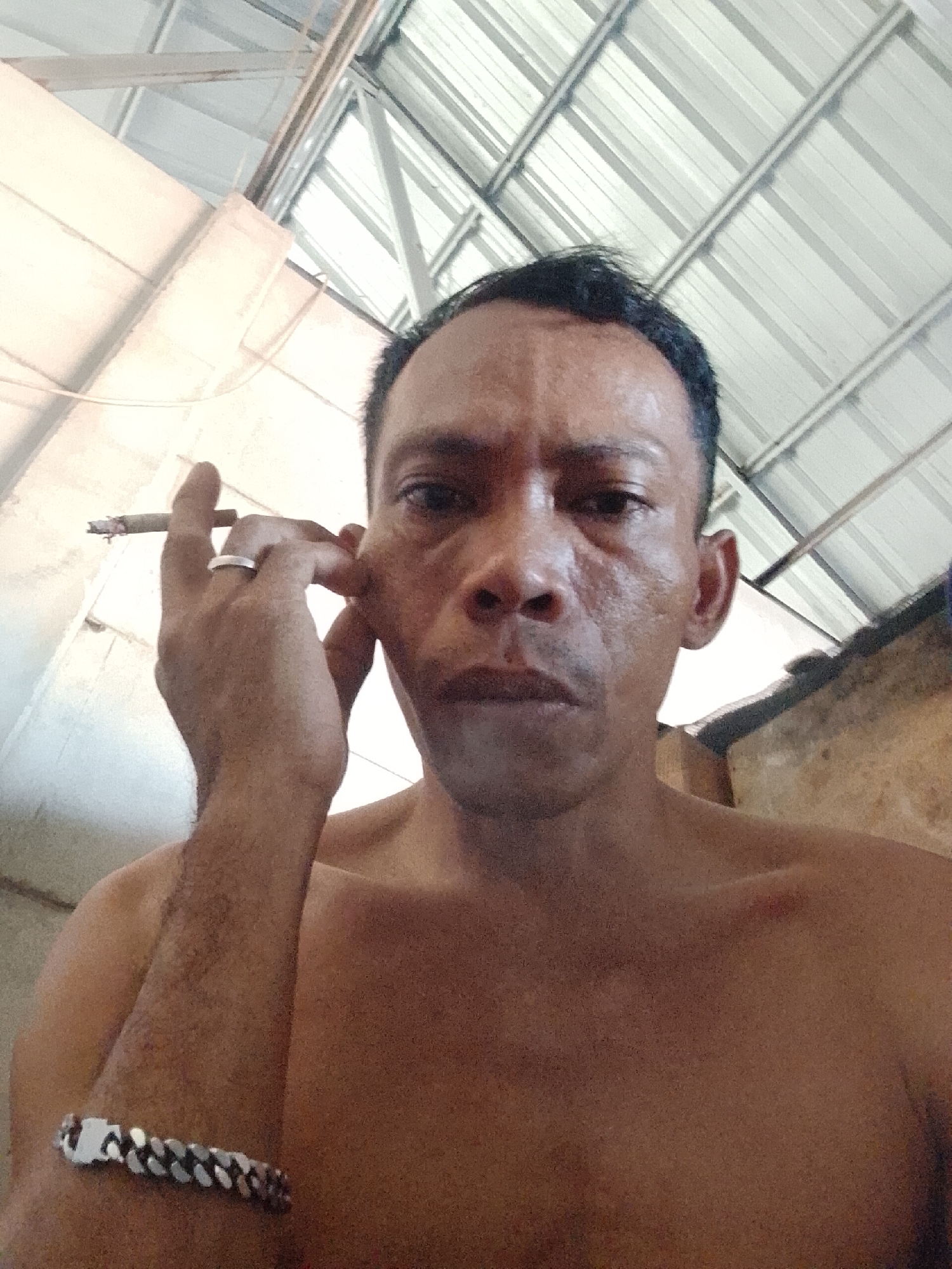 A close-up, low-angle shot depicts a bare-chested man holding a lit cigarette between his fingers, his hand near his face. He wears a ring on his finger and a bracelet on his wrist. The setting is indoors, under a corrugated metal roof. The location is Bukit Ketok, Indonesia.