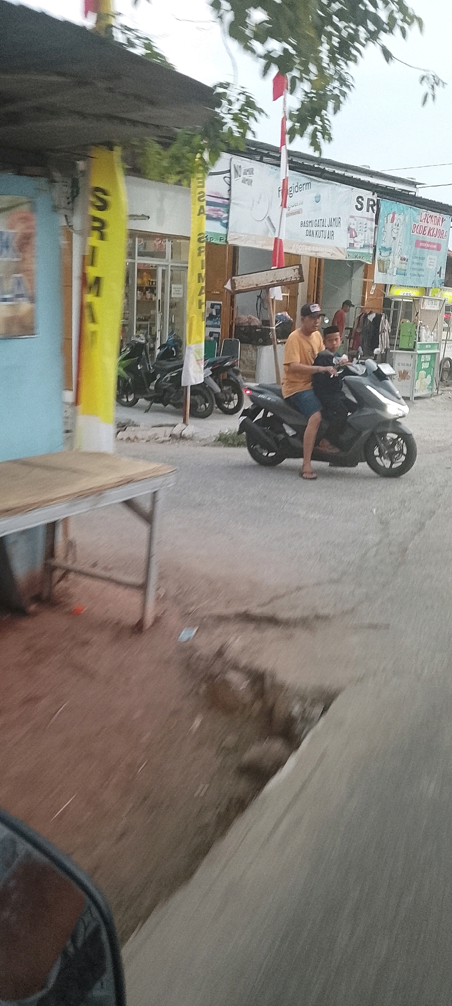 Two individuals, an adult male and a child, ride a dark gray scooter on a paved road in Srimahi, Indonesia. The adult male, wearing an orange t-shirt and blue shorts, drives the scooter while the child, wearing a dark shirt and cap, sits behind him. The scooter has illuminated headlights. In the background, there are shops and stalls, with motorcycles parked in front of them. Red and white Indonesian flags are visible on poles. The scene is captured from a low angle, possibly from another vehicle, and there's a blur effect due to motion.