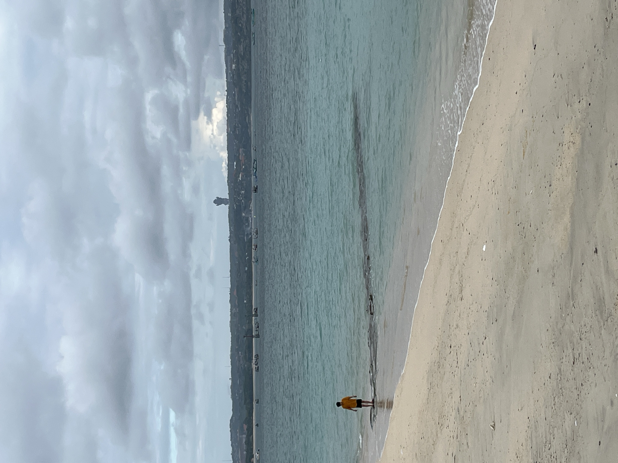 An overcast day at a sandy beach in Kuta, Indonesia. A lone figure, wearing a yellow shirt and black shorts, stands on a shallow rocky outcrop in the turquoise water near the shoreline. Several boats are visible in the distance on the sea, and the Kuta skyline stretches across the horizon. A large, dark statue or monument is prominent against the cloudy sky. The sand is light brown with scattered debris.