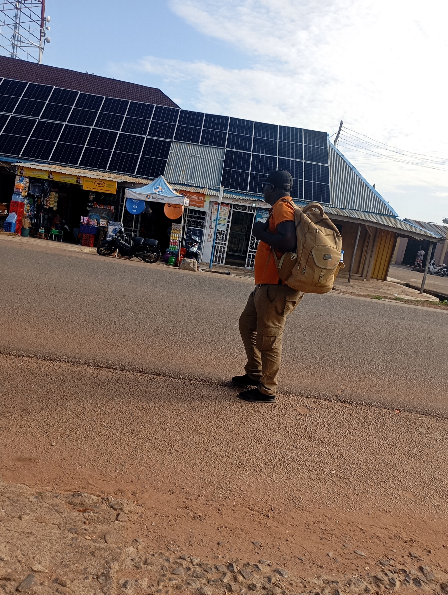 A man wearing an orange t-shirt and brown cargo pants is walking with a large tan backpack on his shoulders on a paved road in front of shops in Shendam, Nigeria. The shops have corrugated metal roofs and awnings. One shop has solar panels installed on its roof. There are motorcycles parked in front of some of the shops, and various items are displayed for sale. The sky is partly cloudy.