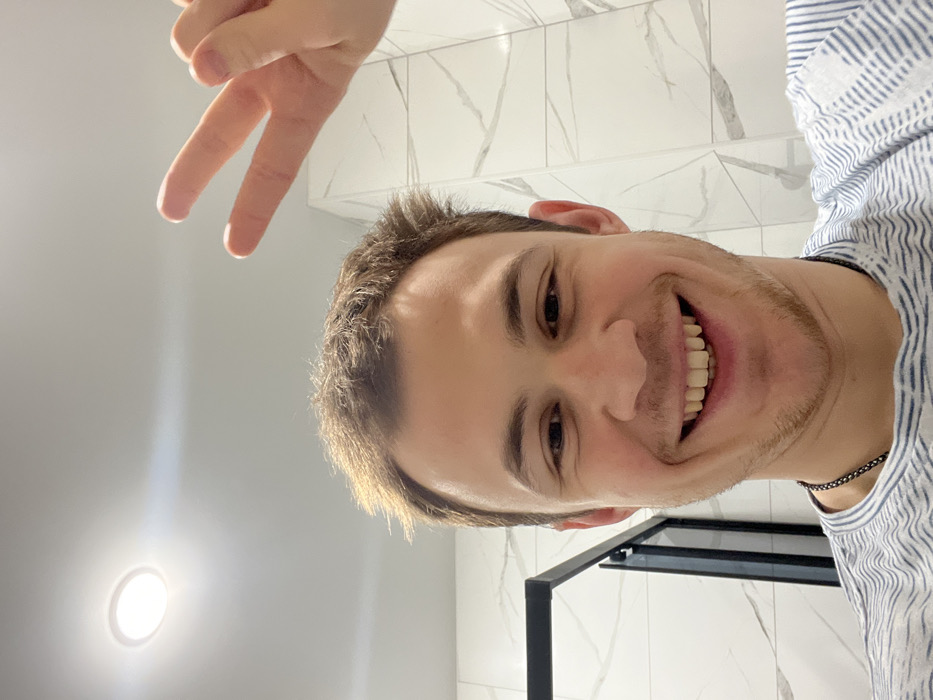 This image captures a close-up selfie of an adult male in an indoor setting, identified as being in Tugurgoi, Russia. The male subject has light skin, short light-brown hair, and light stubble on his jawline and chin. He is smiling widely, revealing his upper and lower teeth, and wears a light-colored t-shirt with a wavy blue and white striped pattern and a dark chain necklace. His right hand is raised, making a "peace sign" (V-sign) with the index and middle fingers extended, positioned in the upper right quadrant of the frame. The background consists of white rectangular wall tiles with a subtle grey marble-like veining. A black matte metal frame, possibly part of a shower enclosure or door, is visible on the left side. The white ceiling features a single bright, circular recessed light fixture in the upper left.
