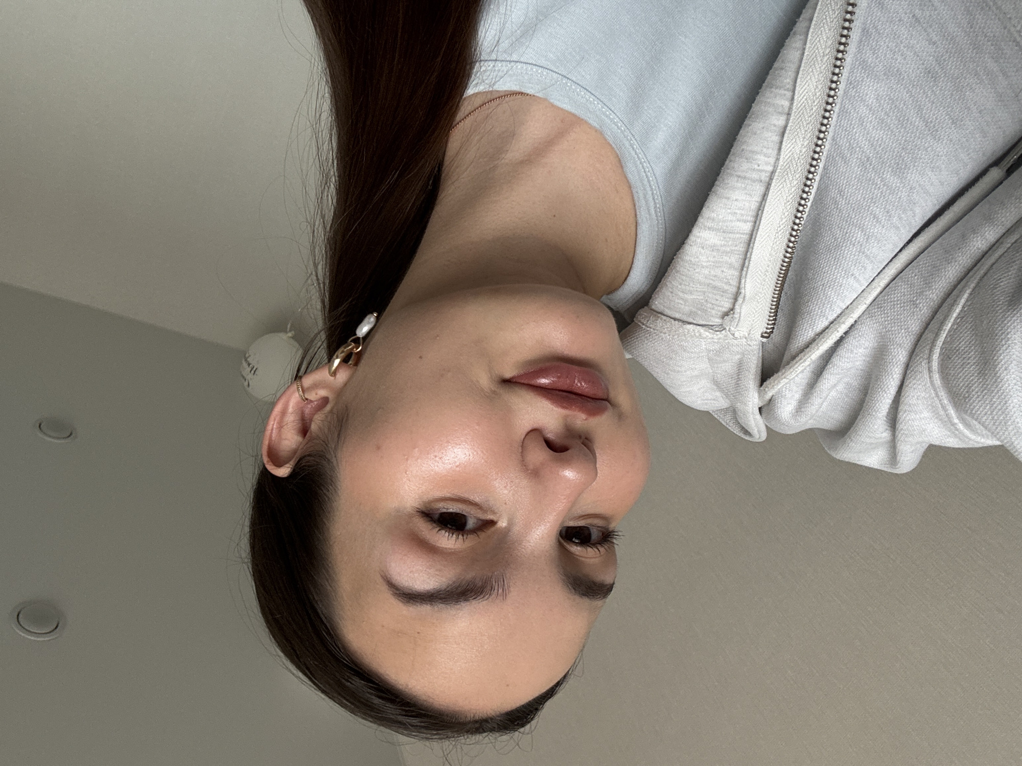 An inverted portrait features a young woman, likely in Chaikovsky, Russia, captured from a low angle looking up towards a light-colored ceiling. Her head is positioned at the bottom of the frame, with long, dark brown hair flowing upwards. Her face presents a luminous, hydrated complexion, with full eyebrows, dark eyes looking downwards, and glossy, rose-tinted lips. She is dressed in a light grey zip-up hoodie over a lighter blue t-shirt.
In her visible ear (her left ear, appearing on the viewer's right), she wears a small gold hoop in the upper cartilage and a larger gold hoop with a white, irregular pearl-like drop in the lobe. A gold ring is also visible on the ring finger of her left hand (appearing on the viewer's right due to the image's inversion). The background shows two recessed circular light fixtures on the ceiling and a partially visible white, cylindrical object. The woman holds a calm, neutral expression, looking towards the camera. The indoor setting suggests ambient artificial lighting. No visible text is present.
