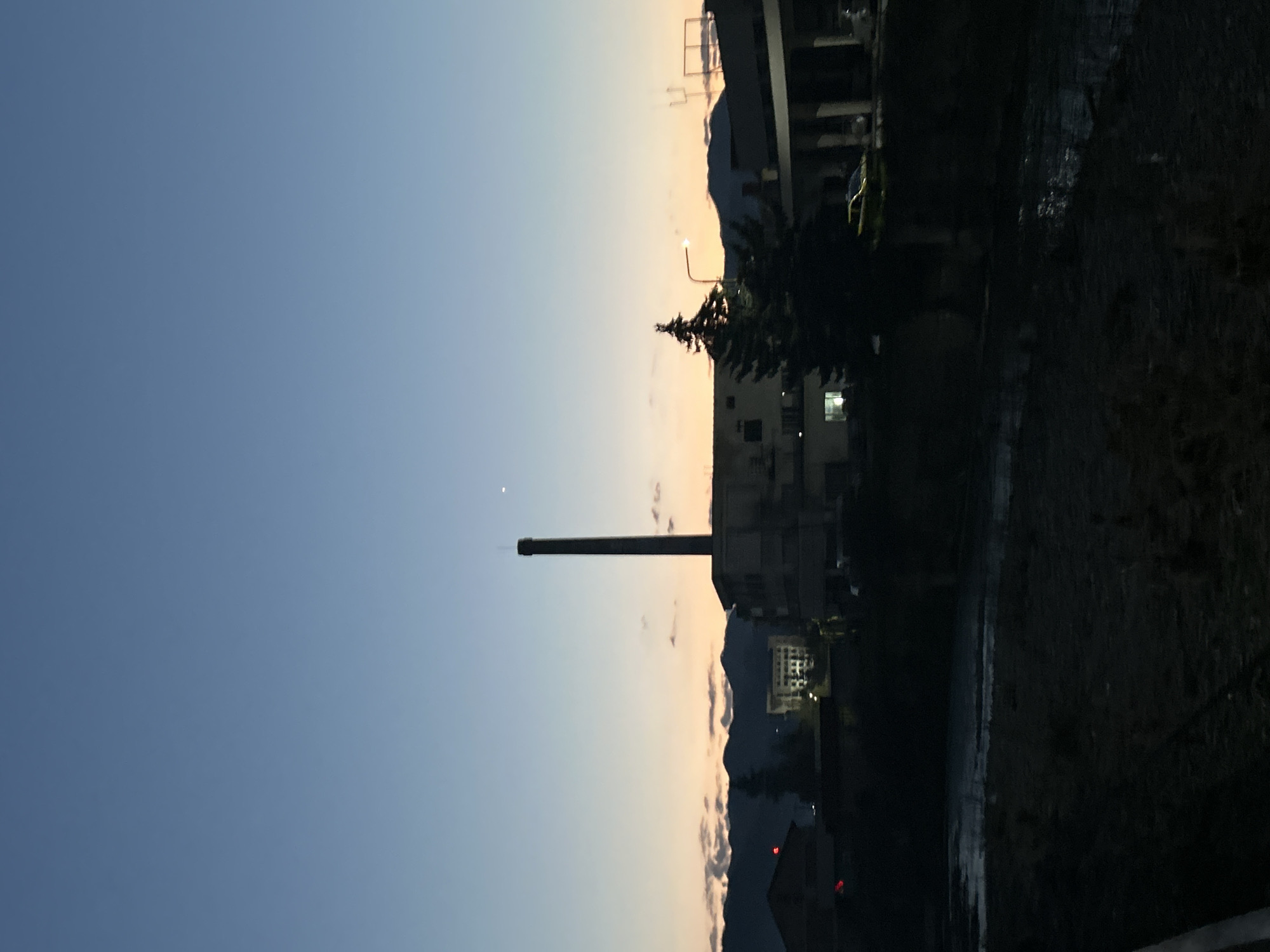 The image captures an evening scene in Zlatograd, Bulgaria, at dusk. A tall, dark smokestack stands prominently against the gradient of the sky, transitioning from a pale orange near the horizon to a deep blue overhead. The buildings are partially obscured by the dim light, and mountains are visible in the background. A street light emits a soft glow near the tree. The foreground shows a dark, textured surface.