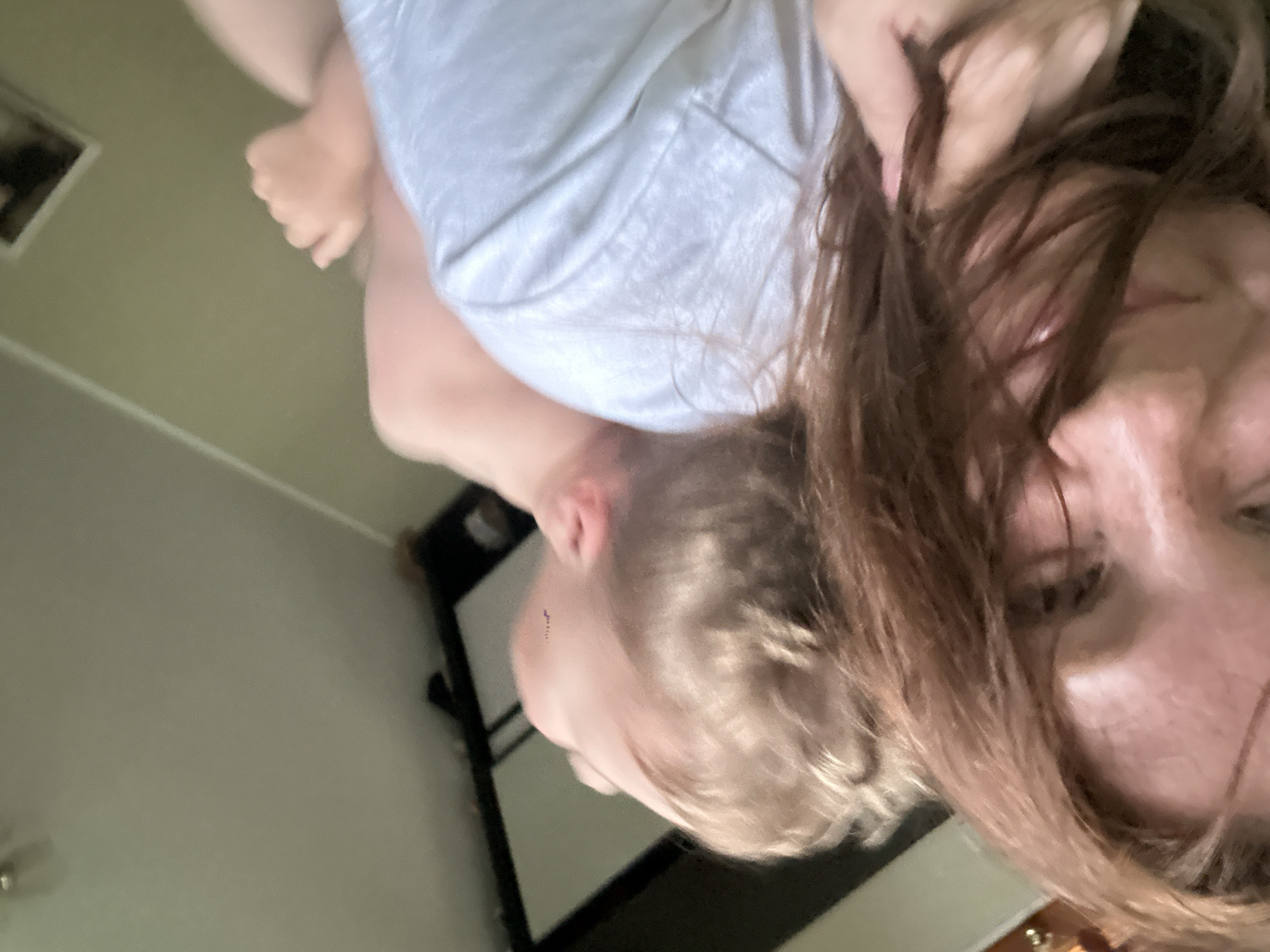 A woman with long brown hair is holding a child with blond hair and a white shirt. The scene appears to be indoors. A bed or headboard is partially visible in the background. The orientation of the image is rotated.