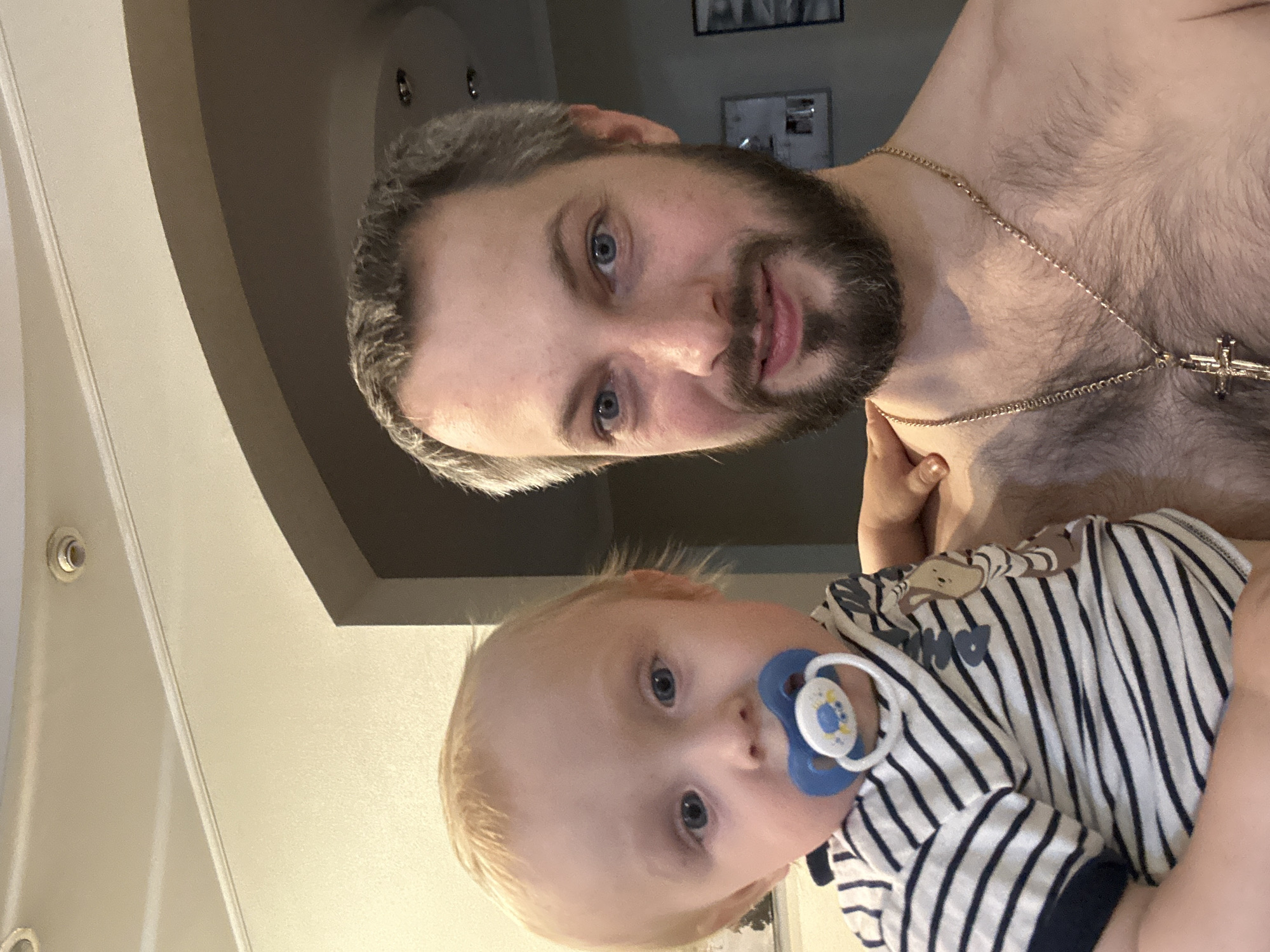 A man with a beard and graying hair, wearing a gold chain with a cross, holds a baby with a pacifier. The baby is wearing a striped shirt. They are indoors, with a ceiling and an arched doorway visible in the background.