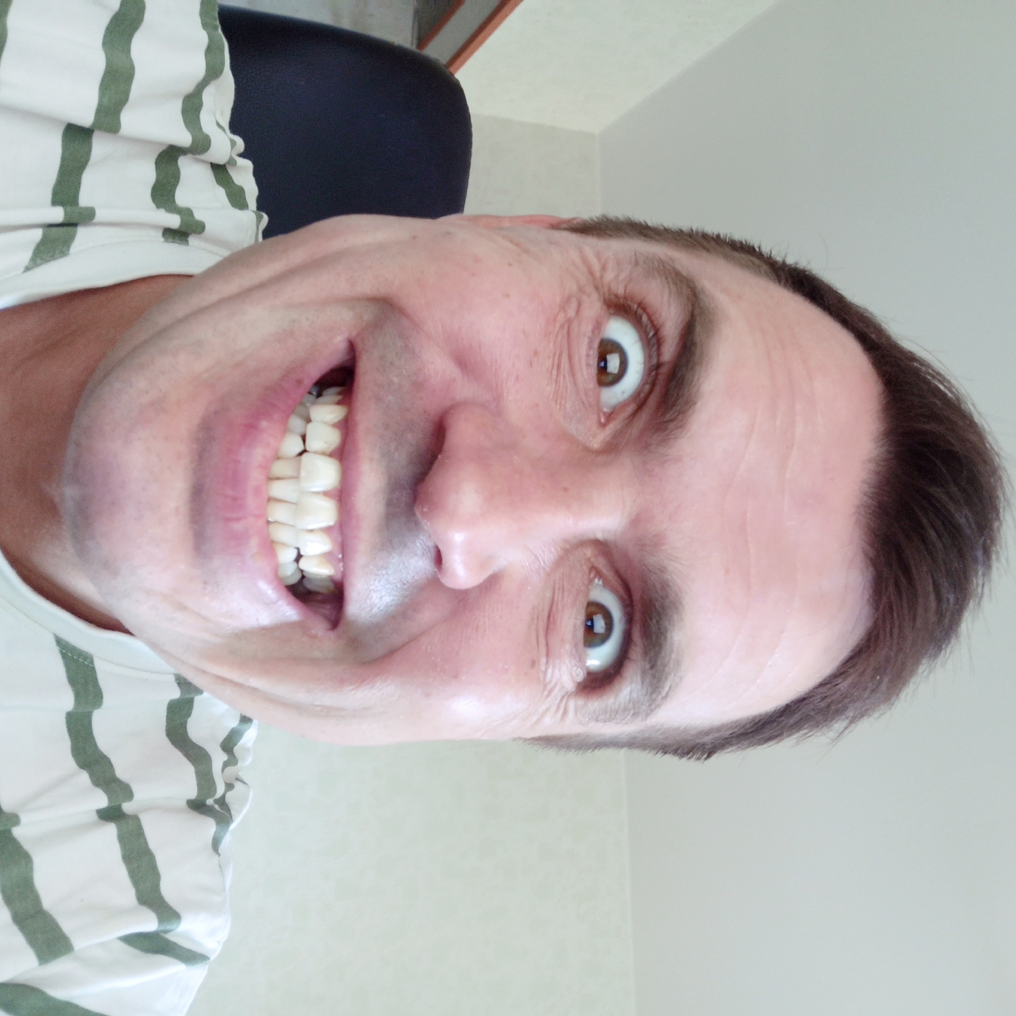 The image shows a close-up, slightly distorted, selfie-style photograph of a man's face. He is looking directly at the camera with his eyes wide open and a wide smile, revealing his teeth. The man has brown hair and is wearing a white t-shirt with green horizontal stripes. His left shoulder and part of a dark chair are visible behind him. The background appears to be an indoor setting, possibly a room with light-colored walls. No specific location in Nizhny Novgorod, Russia, can be identified from the image.