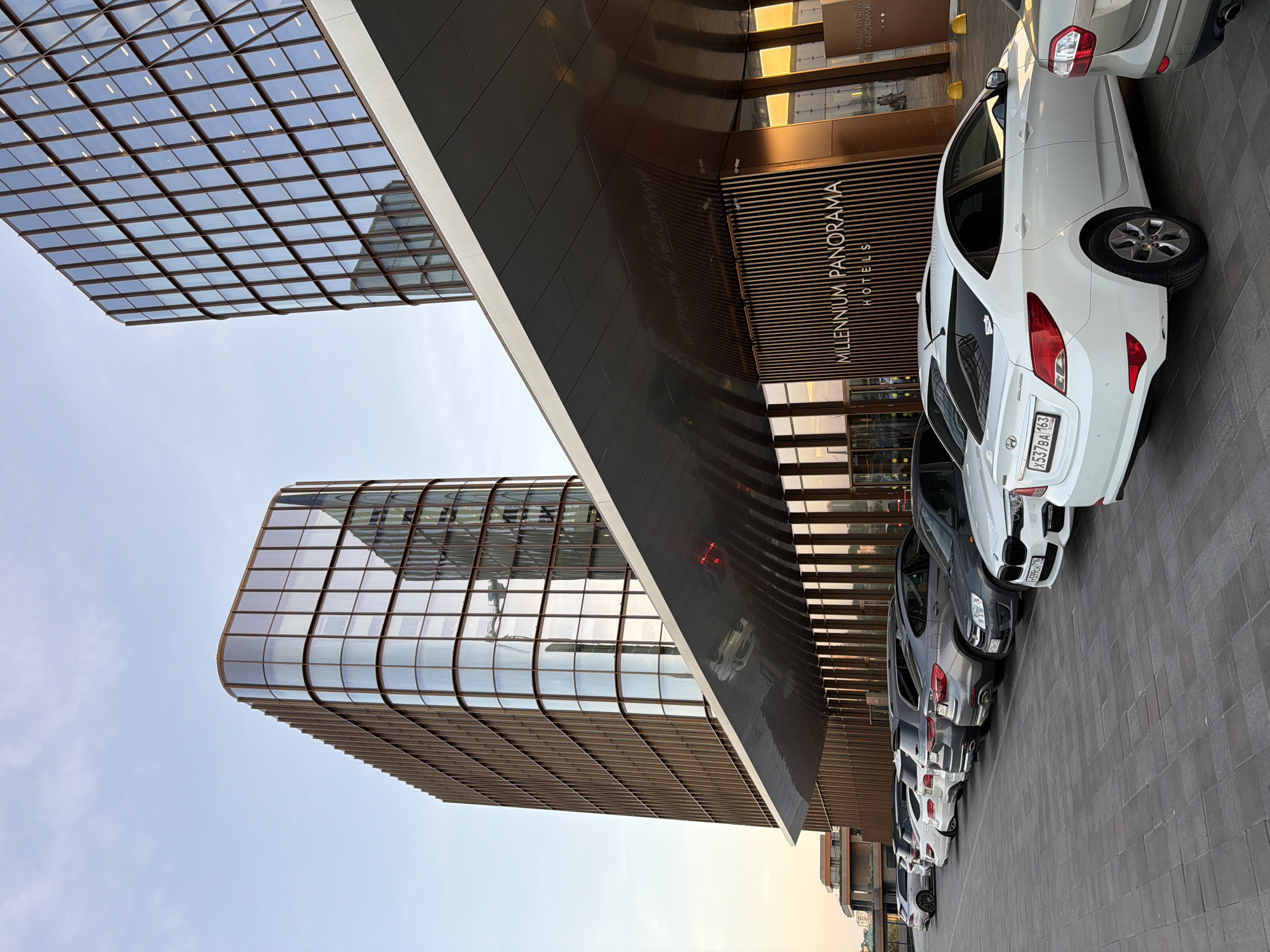 The image shows the exterior of the Millennium Panorama Hotel in Kazan, Russia. The building features modern architecture with glass and metal panels. Several cars are parked along the side of the hotel. The sky is visible above the buildings, suggesting it is daytime.