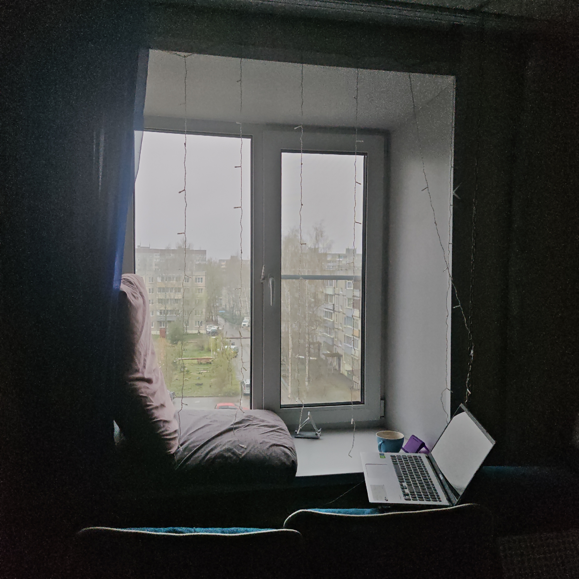 The media file displays an indoor scene looking out a large window, creating a striking contrast between the dimly lit interior and the brighter exterior. No people are visible.
The window, framed by dark walls and a dark blue curtain on the left, is adorned with vertical strings of small, delicate fairy lights. A wide windowsill serves as a functional space. On the left side of the sill, a large, rumpled grey-purple cushion is positioned for seating. On the right, a silver laptop with its screen open and brightly lit is placed, accompanied by a blue mug and a small, purple, cube-like object. A small, silver stand (possibly for a phone) is also on the sill.
Beyond the window, an urban landscape unfolds under an overcast sky, indicating daytime. The view consists of several multi-story apartment buildings, typical of post-Soviet architecture, with some trees showing early signs of spring foliage. A road with a parked red car and a grassy area are also visible.
The overall scene suggests a cozy and functional window-side nook, possibly for work, study, or relaxation. The overcast weather contributes to a soft, diffused light outside. The architectural style of the buildings provides a strong cue for the location being Kladovitsy, Russia.
A sticker on the laptop's palm rest is faintly visible, bearing the text "Intel Iris Xe Graphics."