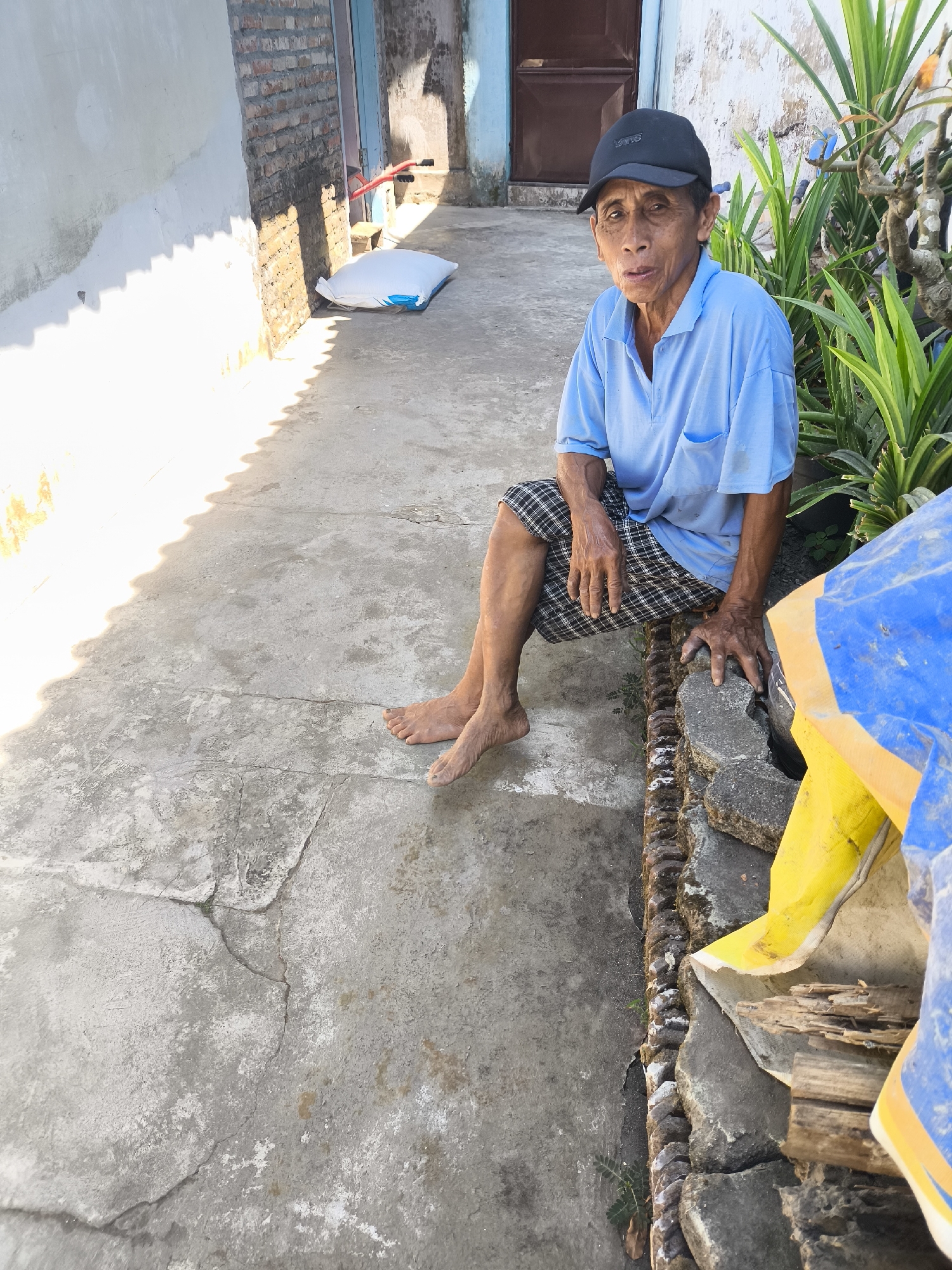 An elderly Indonesian man wearing a blue polo shirt and a black baseball cap sits on a low, rocky border beside a concrete pathway. He is looking directly at the camera with a slightly open mouth. His legs are crossed, and he is barefoot. To his left, a white sack and part of a red wheeled object are visible against a brick wall. To his right, green plants and a large blue and yellow tarp are present. The scene appears to be an outdoor residential area in Lumajang, Indonesia, with a building and a wall in the background. The lighting suggests it is daytime, with strong shadows cast on the concrete.