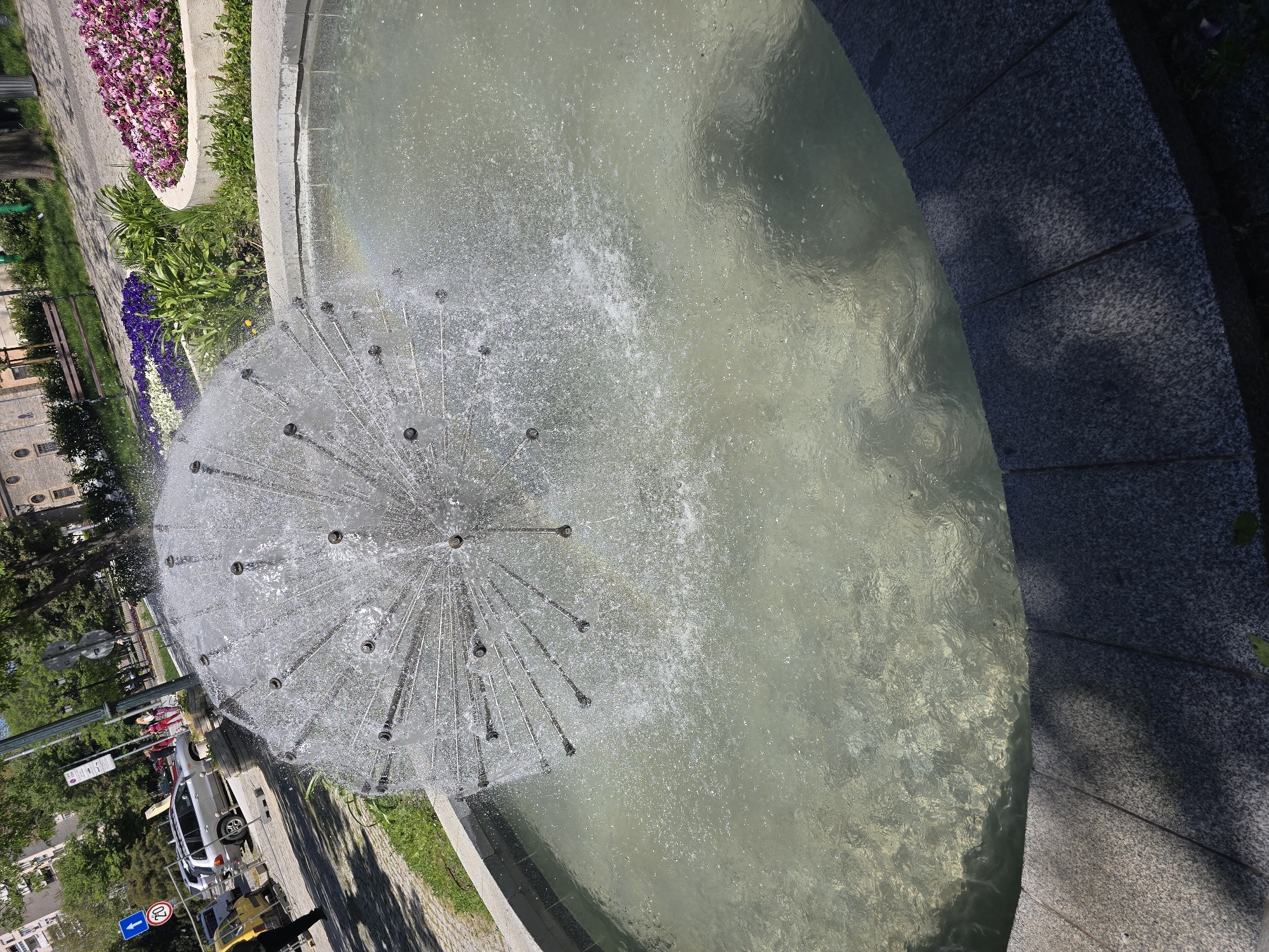 A high-angle, slightly tilted shot reveals a fountain in a city. The fountain has a spherical spray nozzle, and the water glistens in the daylight. The fountain is surrounded by a concrete rim and a flowerbed with colorful flowers. A street runs alongside the fountain, with cars, street signs, and pedestrians visible. Buildings and trees line the street. The location is Unknown, Unknown.