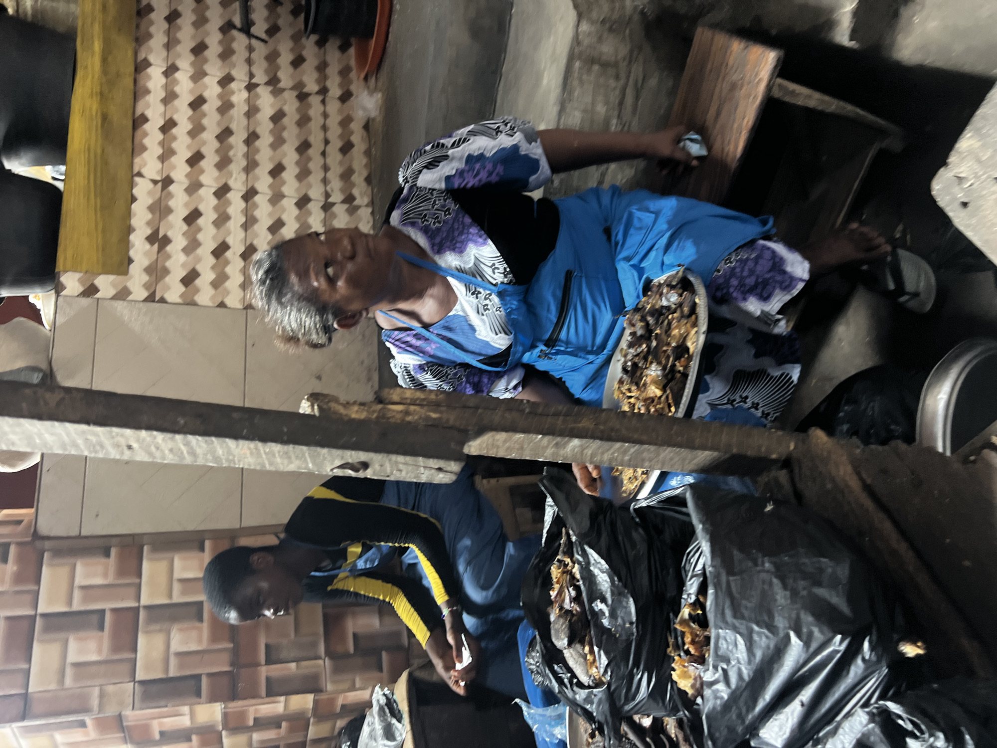 The image depicts a scene in Nkpor, Nigeria, showcasing two individuals involved in the preparation or sale of what appears to be dried fish. An older woman is seated, wearing a blue patterned garment with a black top, and a plate of the dried fish is placed next to her. A younger person, wearing a black and yellow top, is standing. Several black plastic bags filled with the dried fish are visible. The setting seems to be an indoor area, possibly a market stall or a small shop, with tiled walls.