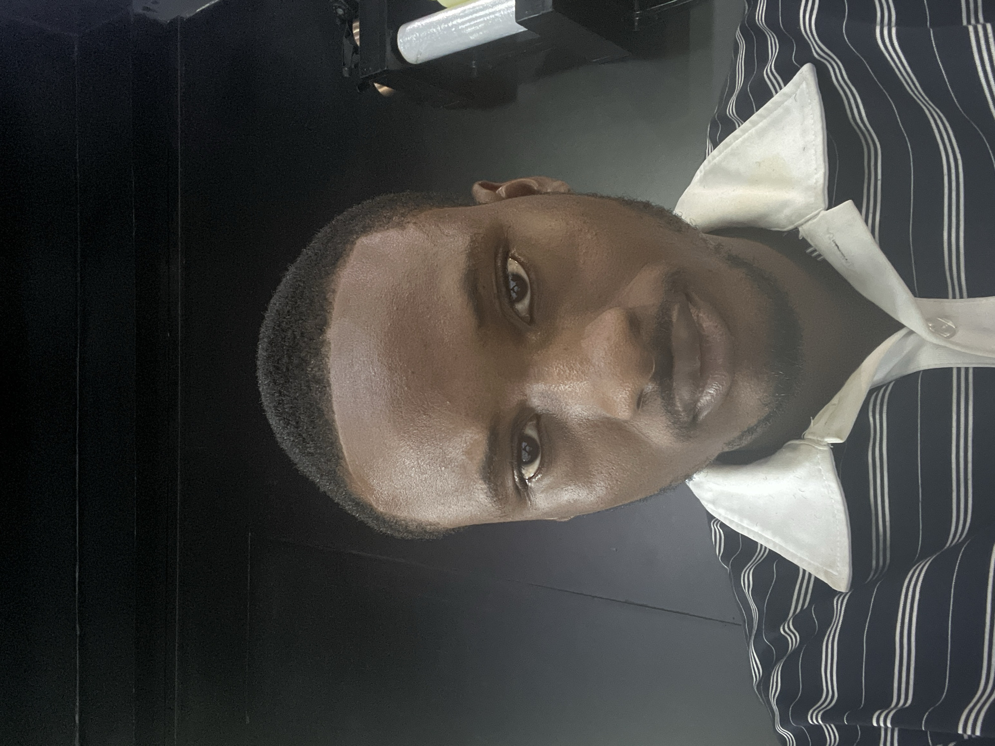 The image shows a man in Lugbe, Nigeria. He has dark skin, short dark hair, and a trimmed beard. He is wearing a vertically striped shirt with a white collar. The background is dark, with some indistinct objects.