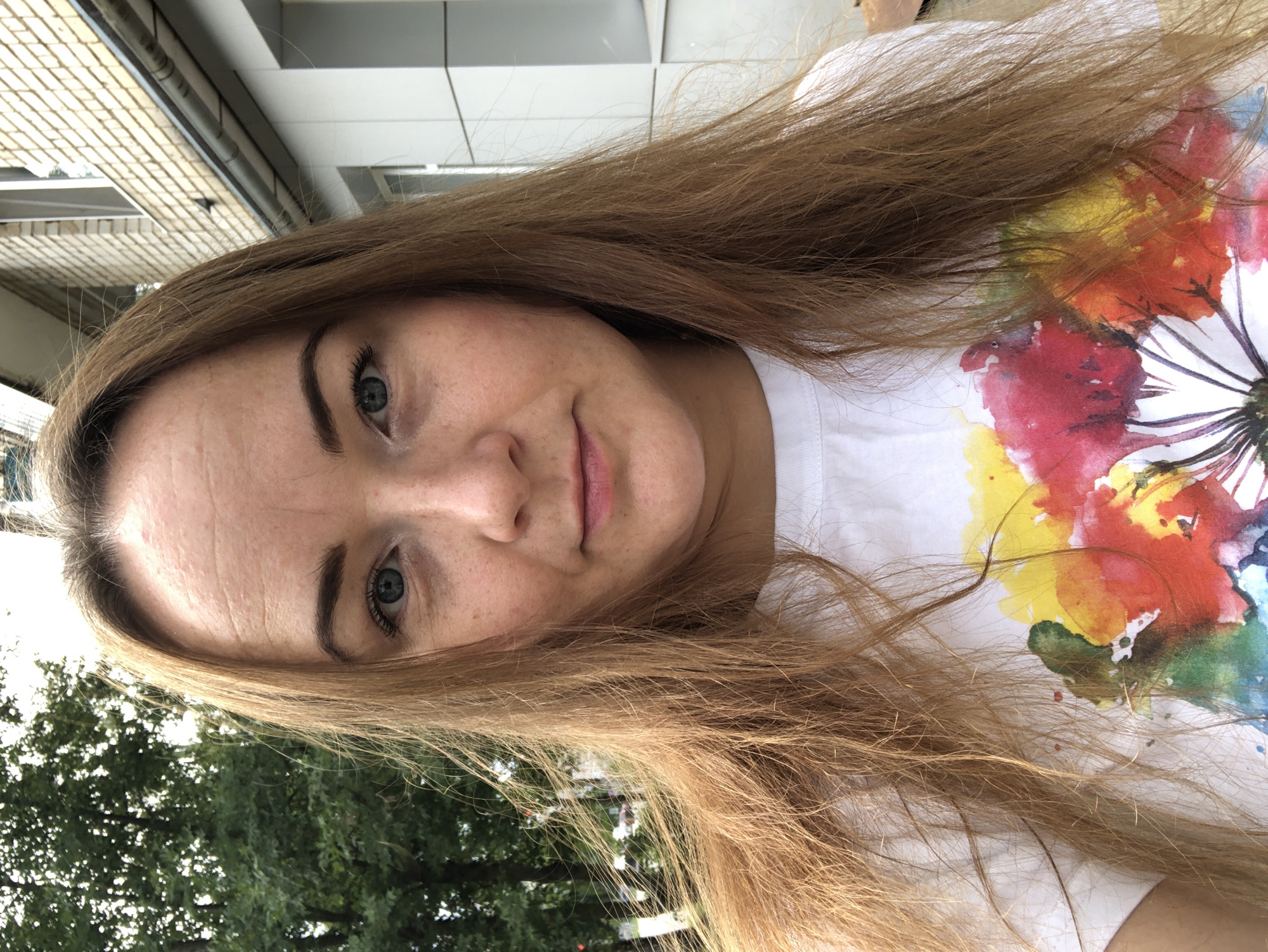 A medium shot shows a fair-skinned woman with long, light brown hair. She has light blue eyes and is wearing a white t-shirt with a colorful floral design. She is outdoors with trees and a building in the background. The building appears to be made of brick and has white trim. The scene is likely in a residential area in Tutaev, Russia.