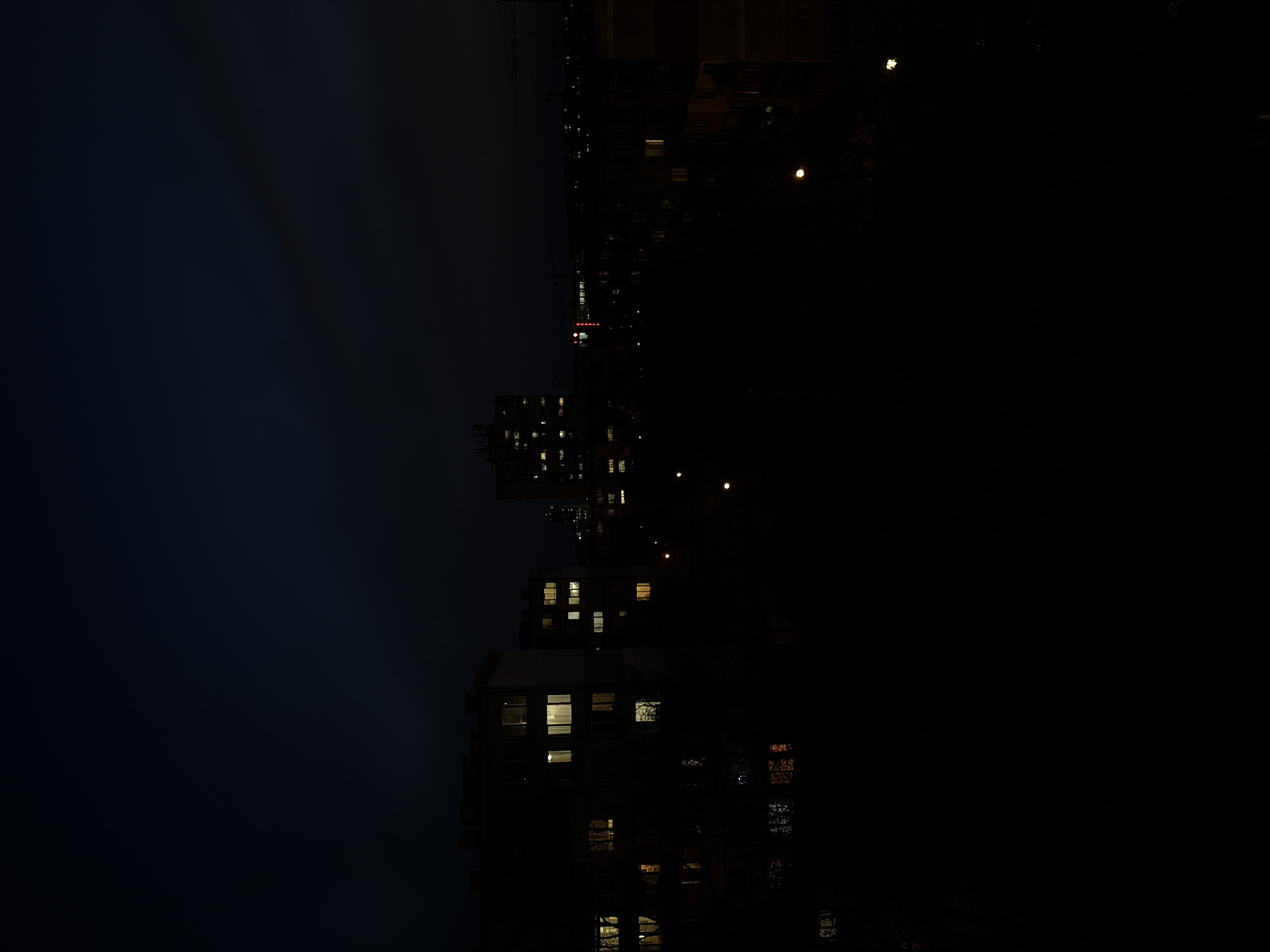 The image shows a nighttime urban scene in Plovdiv, Bulgaria. The view is oriented sideways with buildings illuminated by interior lights standing against a dark blue, almost black sky. The Lincoln building is visible with its name illuminated in red lights. There are street lights. The scene has a generally dark and muted color palette, with the exception of the isolated lights within the buildings and from the street lights.