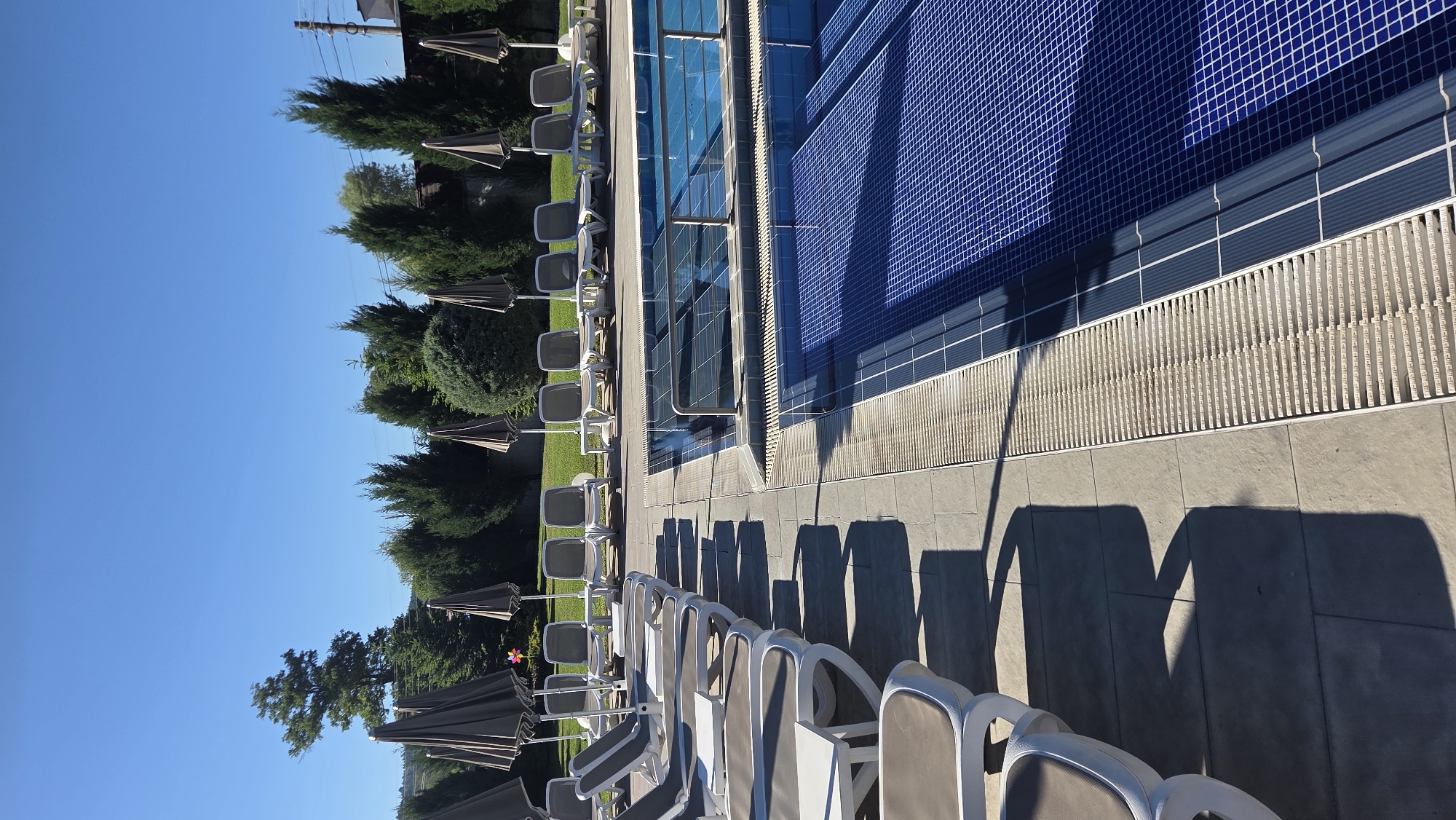 The image shows an empty outdoor swimming pool area on a sunny day. The pool is tiled in blue and has a metal railing. On the left side of the pool, there are rows of empty lounge chairs and closed umbrellas. The ground around the pool is paved with light-colored tiles. In the background, there are lush green trees and a clear blue sky. The scene appears to be a resort or hotel amenity, devoid of any people.