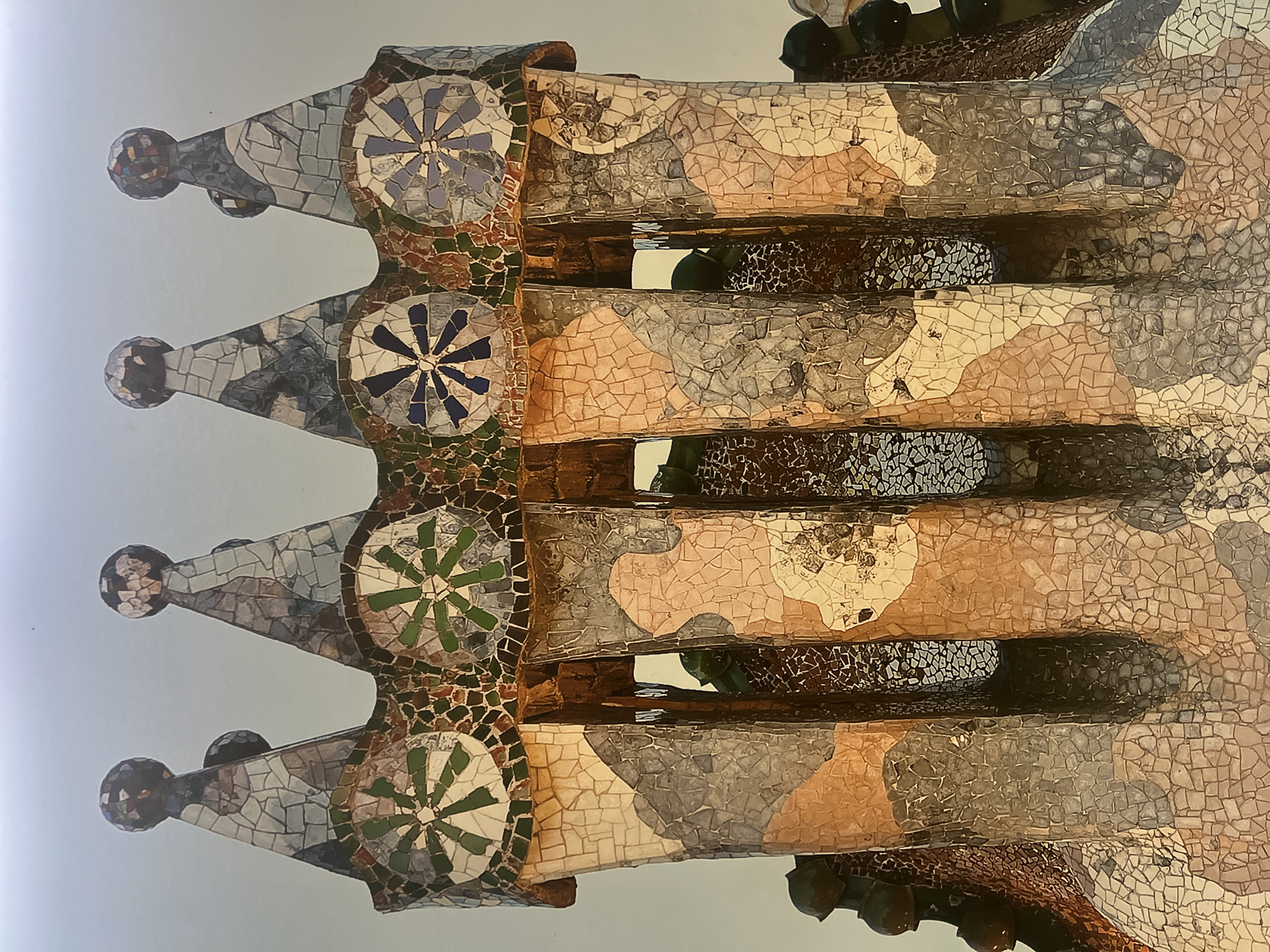 This is a close-up, low-angle shot of the roof chimneys of Casa Batlló, located in Barcelona, Spain. The chimneys are adorned with colorful trencadís mosaics, a technique of broken tile mosaic pioneered by Antoni Gaudí. The mosaics feature a variety of shapes and colors, including white, cream, brown, gray, green, and blue tiles. Each chimney is topped with a conical cap, also covered in mosaics, and some are crowned with spherical finials. The design of the chimneys evokes organic forms, possibly inspired by bones or organic growths. The sky in the background is a pale, hazy blue.
