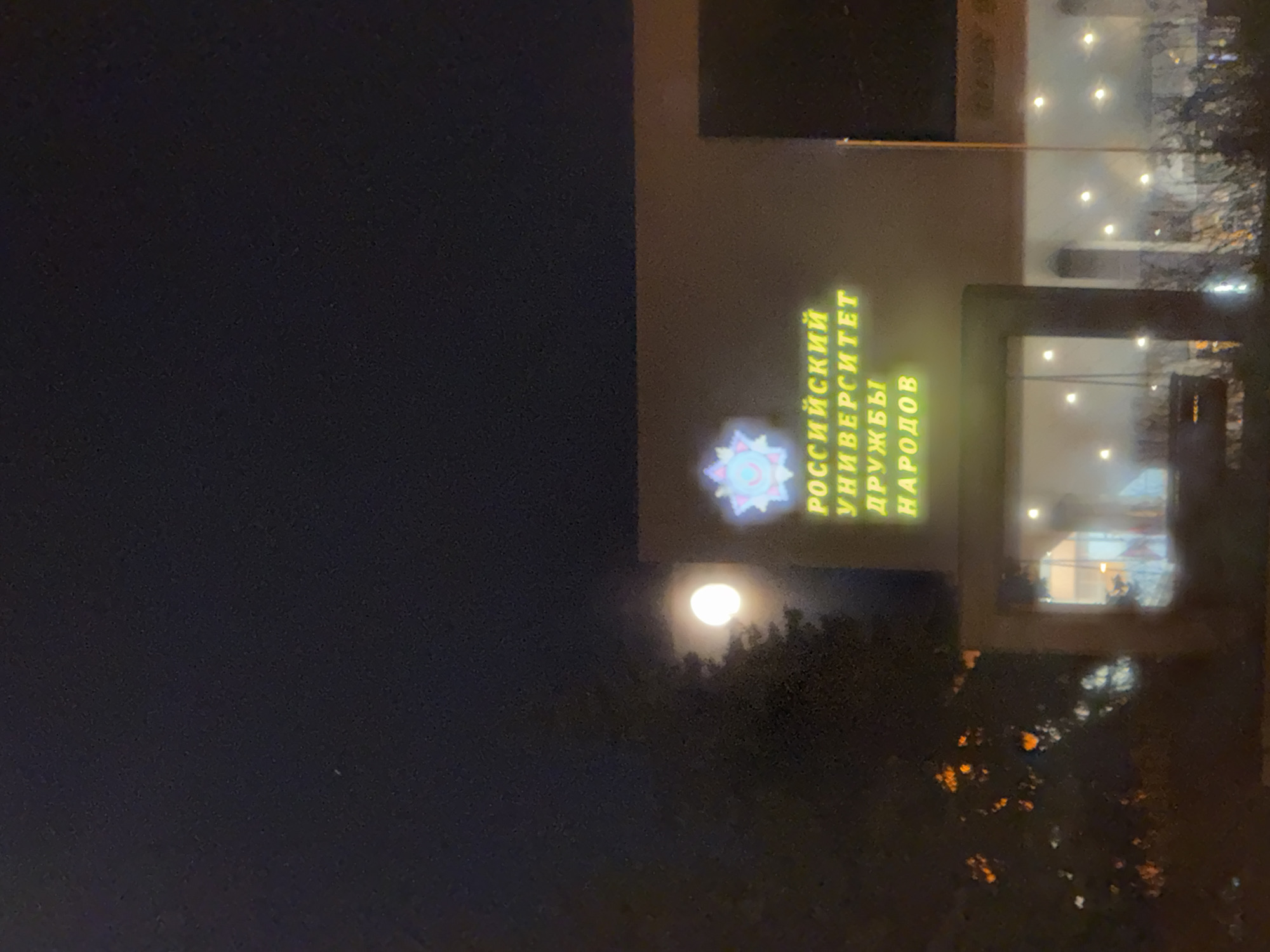 The image shows the exterior of the Peoples' Friendship University of Russia in Moscow at night. Illuminated text and a logo on the building's facade are visible. The full moon is visible in the background of the image. Trees and possibly other vegetation are visible, obscuring the base of the building.