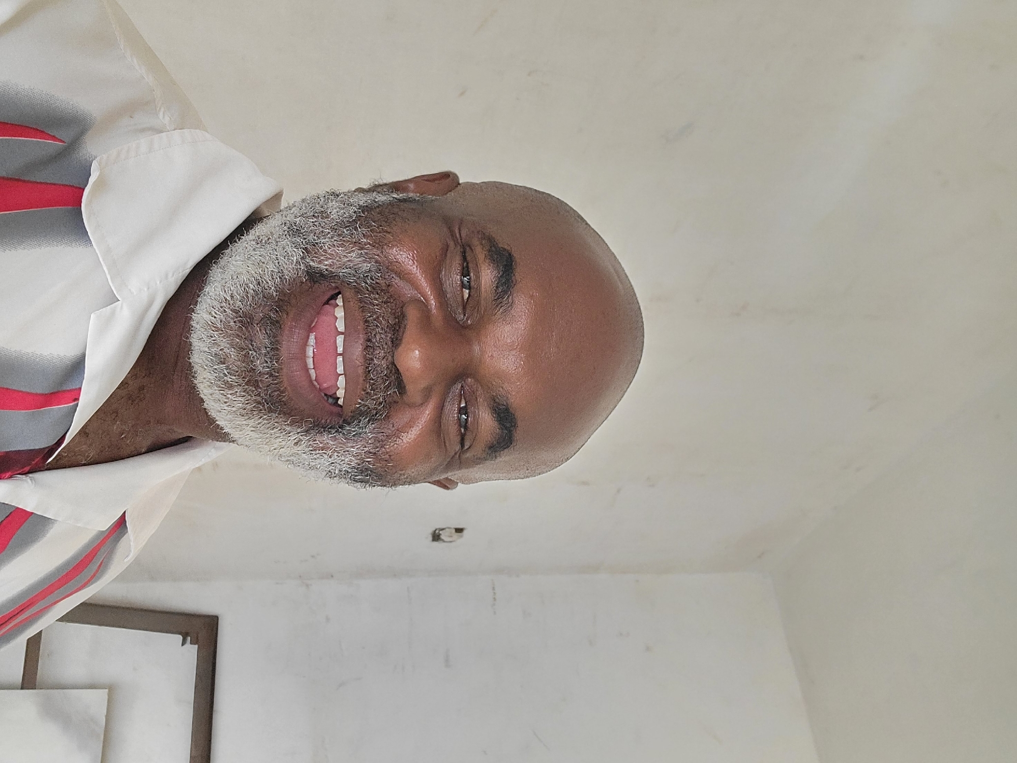 A dark-skinned bald-headed man with a full, mostly-gray beard is smiling at the camera. He is wearing a collared shirt with white, gray, and red stripes. The background consists of a white wall with a framed picture in the lower left corner and a small electrical outlet near the top. The location is Maiburiji, Nigeria.