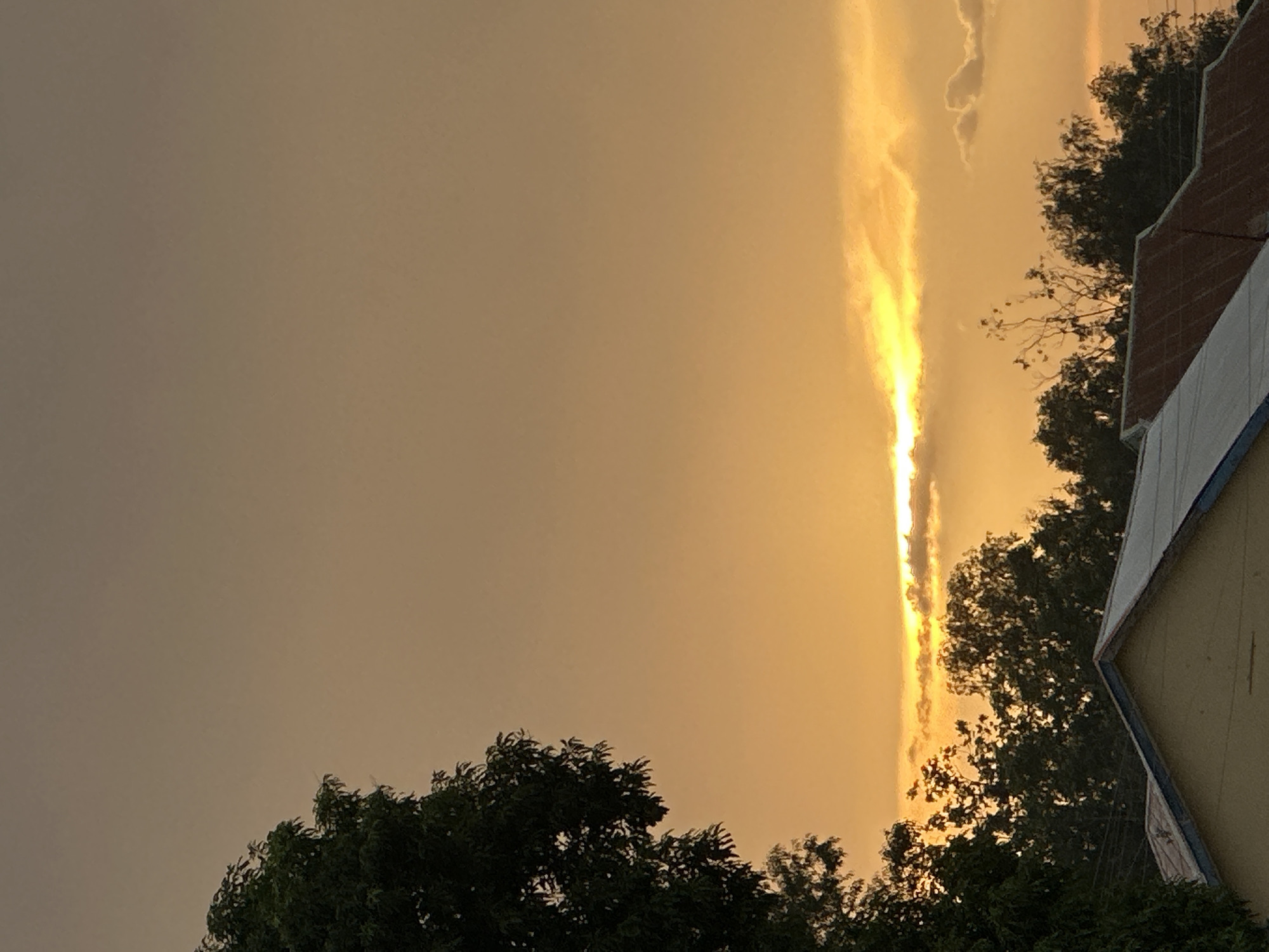The image is a low-angle shot of a sky illuminated by golden light, possibly during a sunset or sunrise. A bright, vertical strip of light cuts through the center of the frame, flanked by darker clouds. Silhouetted trees with dense foliage appear in the foreground, along the bottom of the frame. In the upper right corner, a section of a building is visible, suggesting an urban setting. The location is Bukuru, Nigeria. No people are visible in the scene.