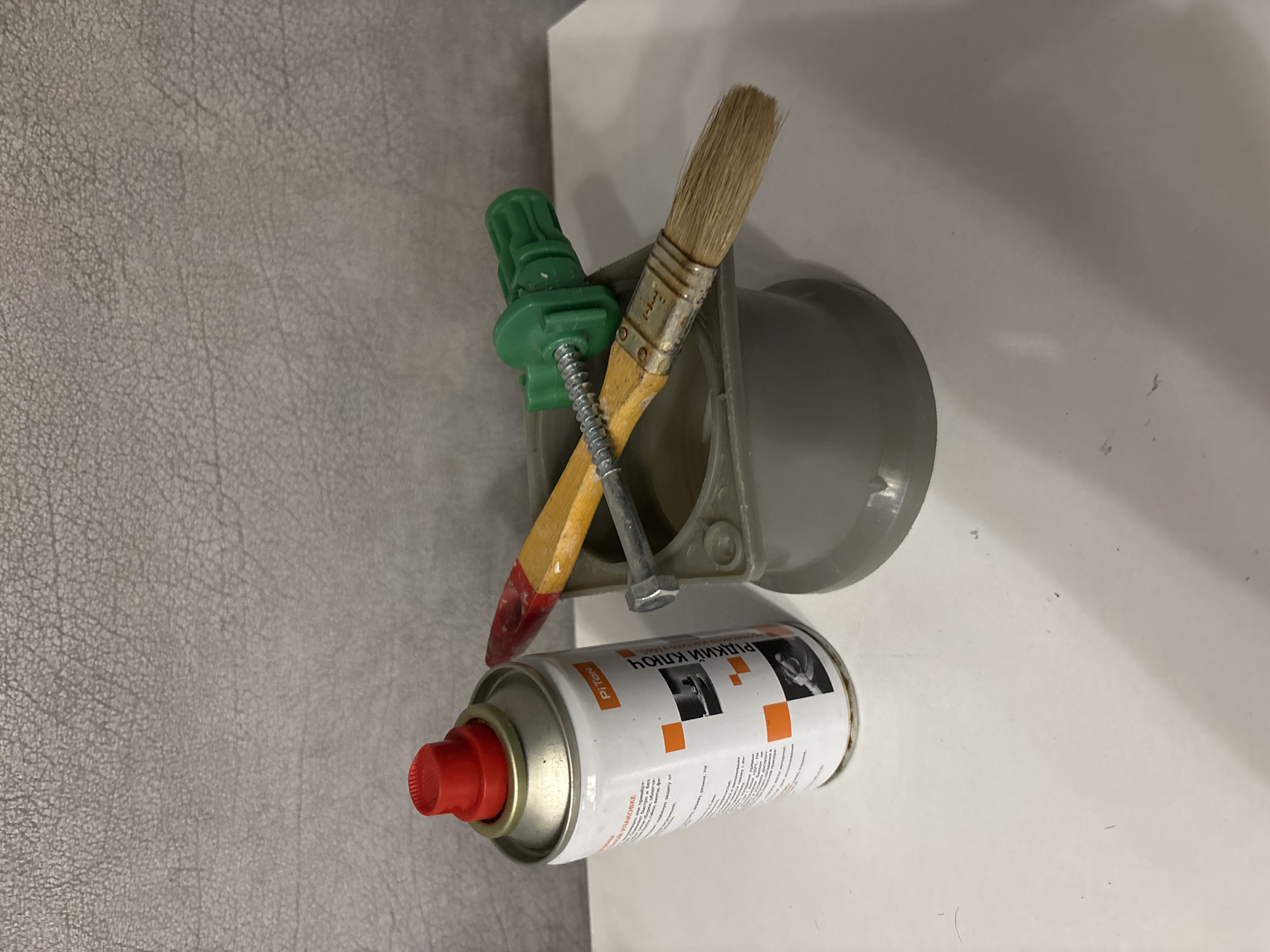The image displays a close-up, rotated view of several DIY tools and a can of spray. In the foreground, a white spray can with a red cap is prominent. The can has text printed on it, including "PITON" and what appears to be Russian lettering that reads "РЫДКИЙ КЛЮЧ". Orange squares are visible on the can's label, along with images of a hand using a tool. Behind the can, a gray plastic object, possibly part of a fixture or housing, is visible. Leaning against this object are two tools: a paintbrush with a wooden handle and metal ferrule, and a pencil-like object with a sharpened, red-tipped end, possibly a crayon or a wax pencil. A green plastic clip or fastener is attached to the pencil. A metal bolt is also present, threaded through the pencil and the green fastener, and extends into the gray plastic object. The background consists of a textured gray surface on the left and a plain white surface on the right, suggesting an indoor setting. The lighting appears to be artificial, casting soft shadows. There are no people or animals in the frame. The overall impression is one of tools laid out, perhaps for a repair or crafting task.