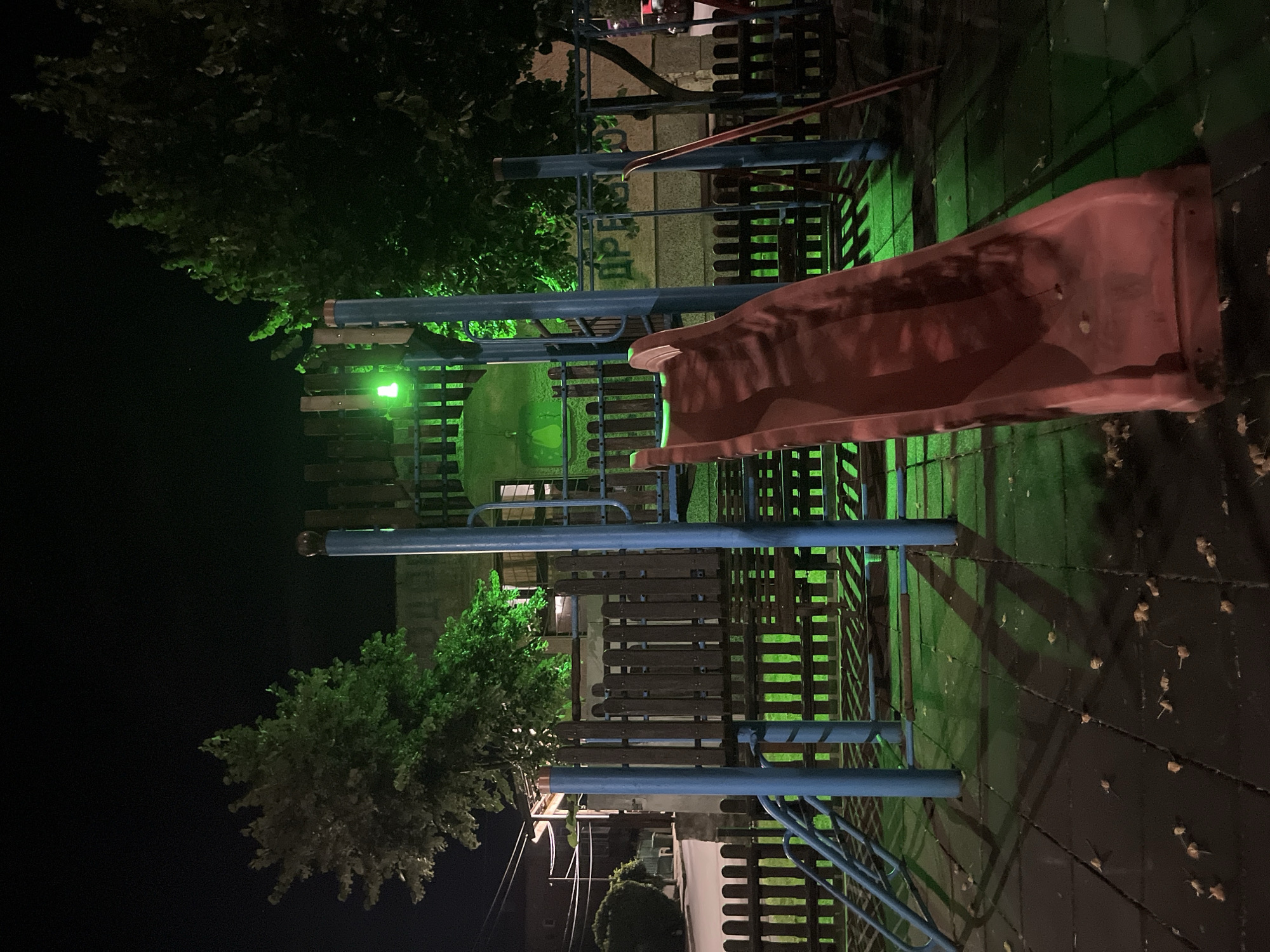 An outdoor playground in Obzor, Bulgaria, is illuminated by green lighting at night. A red plastic slide is the most prominent feature in the foreground, angled towards the right of the frame. Behind the slide is a wooden fence and blue metal support poles for playground equipment. Green light emanates from behind the fence and reflects on the ground, which is a dark surface with scattered fallen leaves. Trees with dark foliage are visible in the background, as is a building. The sky above is dark.