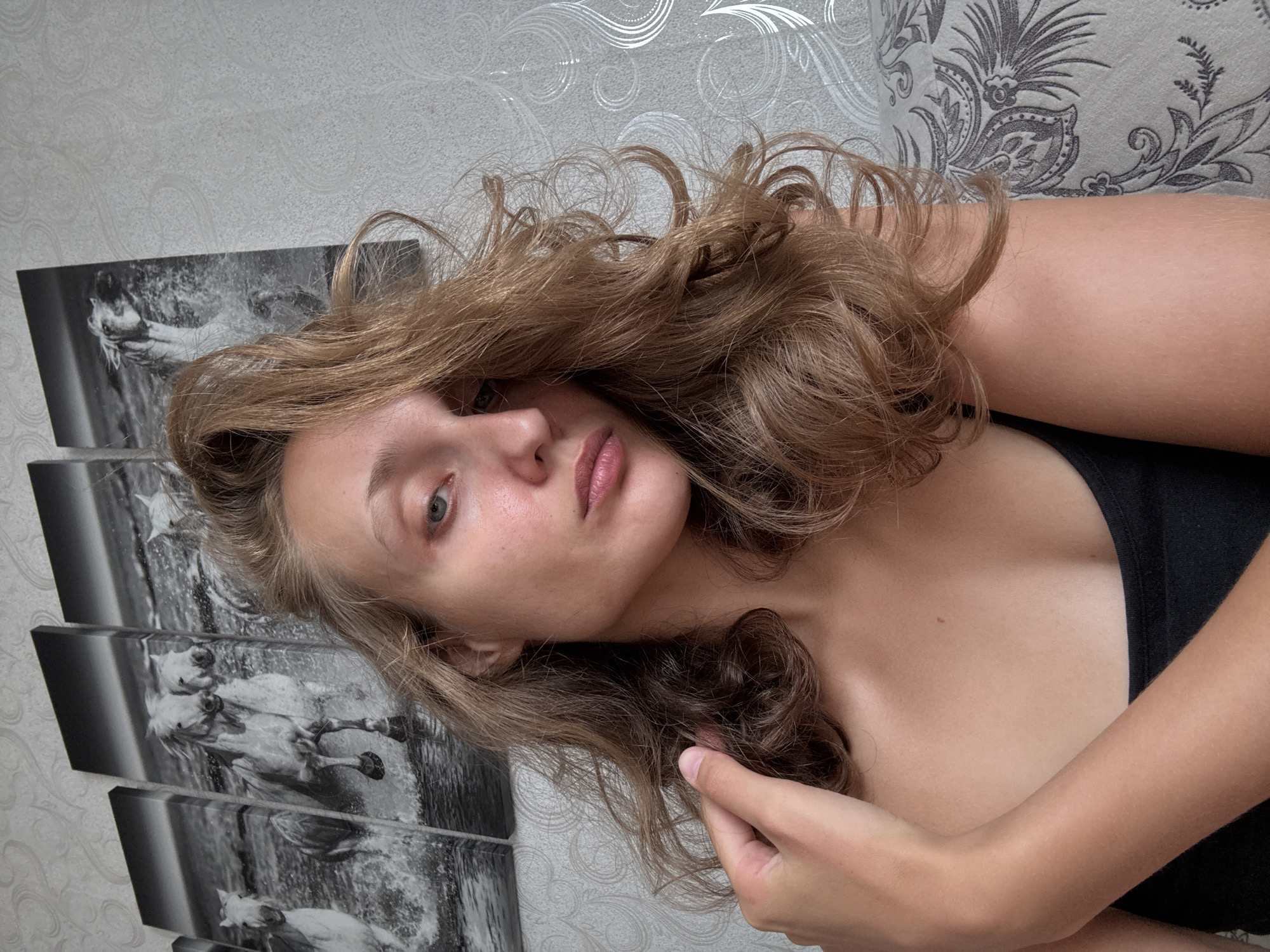 A young woman with long, wavy blonde hair is shown in a close-up selfie. She is wearing a black tank top and her left hand is raised to her hair. The background consists of a textured wallpaper and a triptych wall art depicting white horses running through water. The image is taken indoors, likely in a residential setting, and does not provide any specific clues about the city of Kuyeda, Russia.