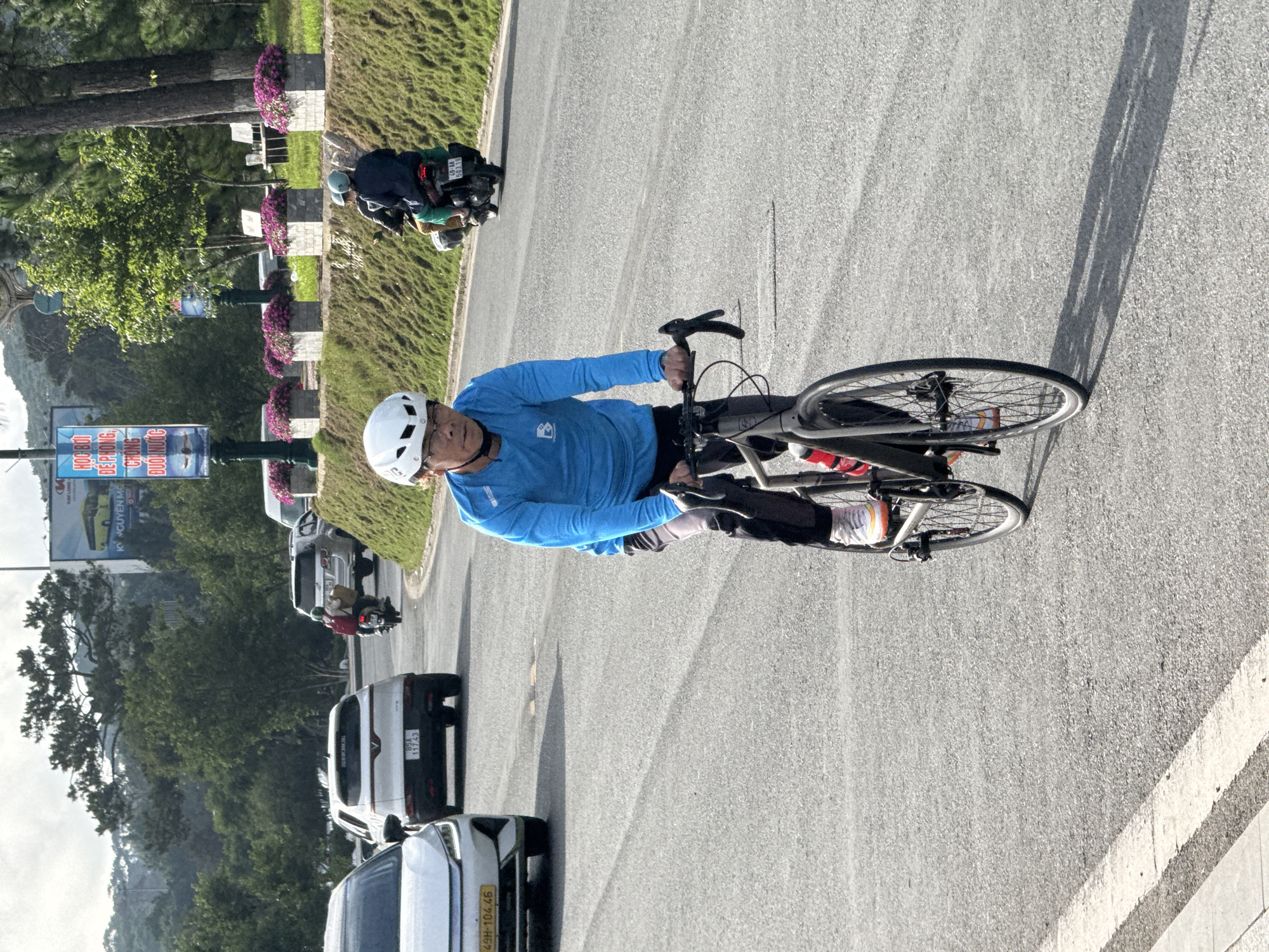 A male cyclist, appearing to be middle-aged, is observed riding a grey road bicycle on a paved asphalt road in Da Lat, Vietnam. He is wearing a white helmet, a long-sleeved blue athletic shirt with a stylized "B" logo, dark pants, and white athletic shoes with orange accents. His hands are on the drop handlebars, and he is looking slightly to his left. A red water bottle is visible in a cage on the bicycle frame. A long shadow from the cyclist and his bike extends onto the road to his right, indicating early morning or late afternoon light.
The road features visible white lane markings on the left. To the cyclist's right, an elevated grassy slope is lined with multiple rectangular concrete planters containing purple flowering plants, with various trees and what appear to be white benches beyond. A motor scooter, carrying two helmeted individuals—the driver and a passenger with a red backpack and a package—is traveling in the same direction as the cyclist further down the road. The scooter's license plate reads "49-AA 503.19". Behind the scooter, a silver SUV is partially visible.
To the cyclist's left, two white VinFast electric vehicles are also traveling in the same direction: a VinFast VF e34 SUV with license plate "85A-117.43" is ahead of a partially visible VinFast VF 8 sedan displaying license plate "49H-104.46".
The background features a densely treed, hilly landscape under an overcast sky. Several large billboards and banners are visible: one prominent billboard shows a yellow bus and Vietnamese text, while a banner on a pole displays "HỌC BƠI ĐỂ PHÒNG, CHỐNG ĐUỐI NƯỚC" (Learn to swim to prevent drowning) and another banner "NGUYÊN NHÂN ĐUỐI NƯỚC ĐIỂN HÌNH" (Typical causes of drowning).