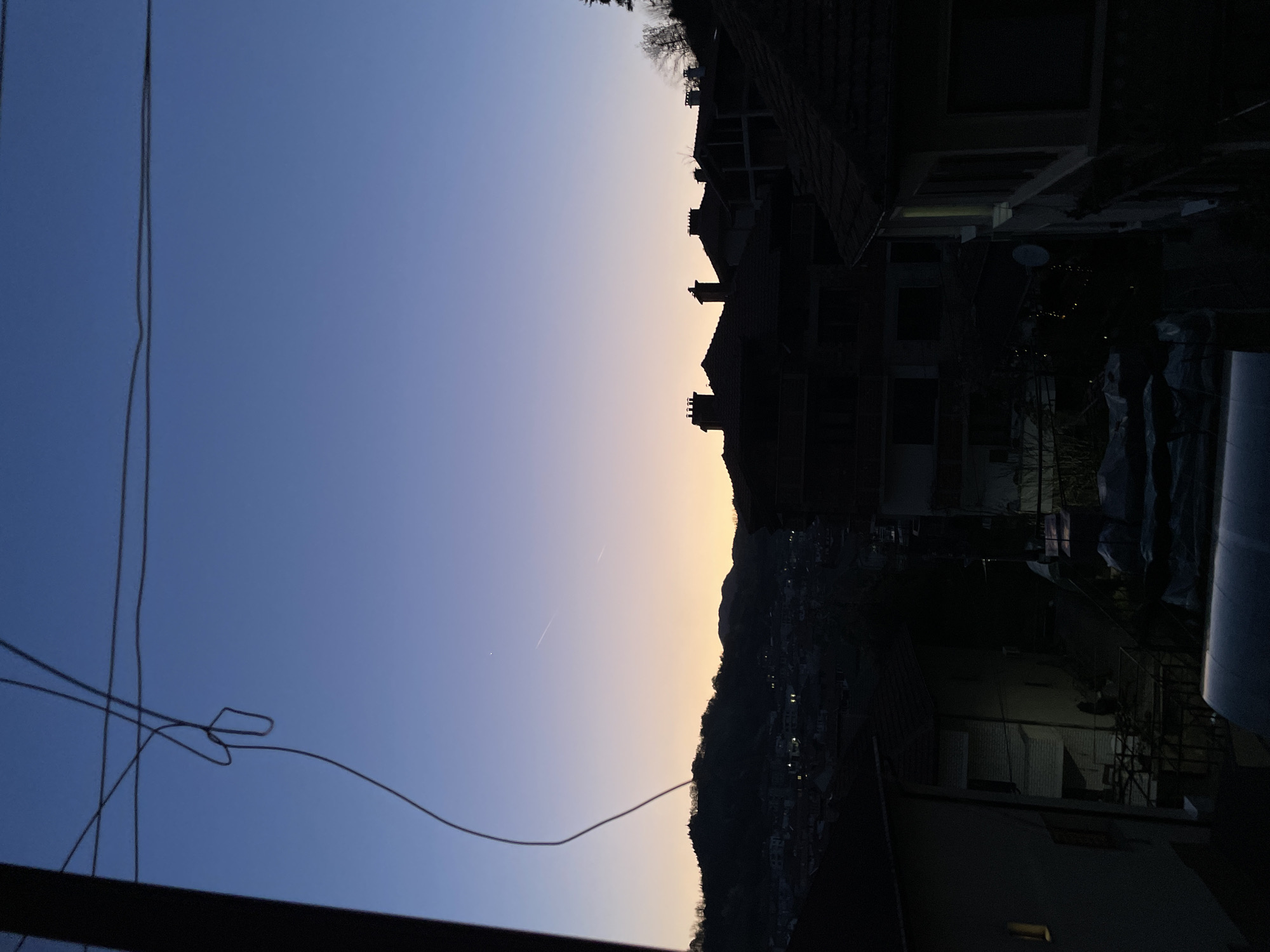 The image shows a view from a balcony in Zlatograd, Bulgaria, at dusk. The sky transitions from dark blue to pale yellow near the horizon. Power lines crisscross the lower left of the frame. Silhouetted buildings with red tile roofs stand on the right. A mountain range with sparse lighting is visible in the distance, separating the sky from the buildings. The foreground is dark.