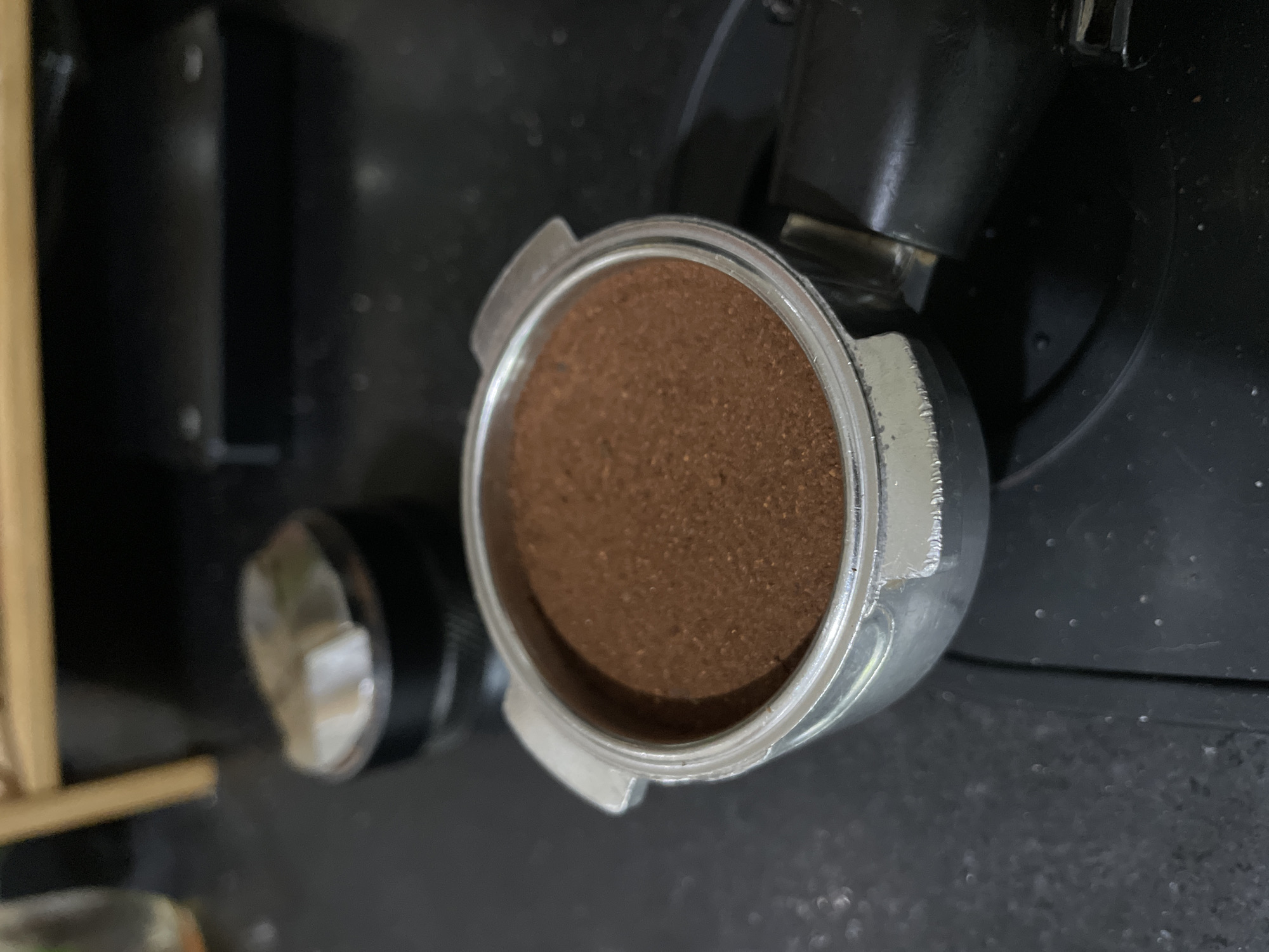 An overhead shot captures a silver-colored portafilter filled with finely ground, dark brown coffee. The coffee grounds appear evenly distributed within the metal basket. The portafilter is positioned on a dark, textured surface, likely a countertop or part of a coffee machine.
In the background, out of focus, are dark, possibly black, components of what appears to be an espresso machine or coffee grinder. To the left of the portafilter, another out-of-focus object with a circular silver/white knob or dial is visible, adjacent to a light-colored, linear object that might be wood or bamboo. The overall setting suggests a coffee preparation area. The lighting highlights the texture of the ground coffee and the metallic sheen of the portafilter. No text is visible in the image.