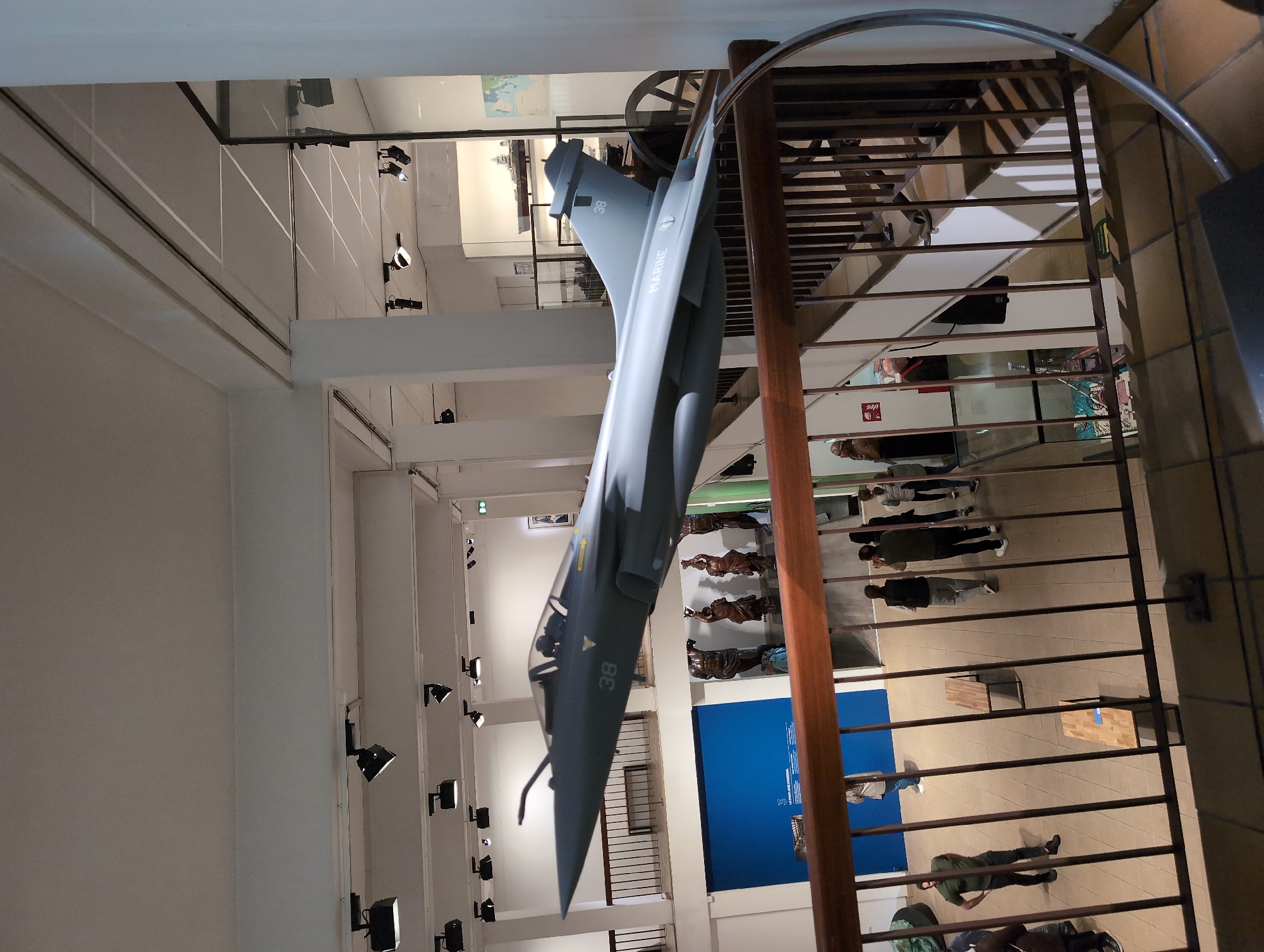 The image captures an indoor scene featuring a suspended Dassault Rafale Marine aircraft model, marked with "38" and "MARINE" on its tail, within a museum-like setting. Viewed from an elevated perspective through a metal railing structure, the exhibition area below displays several people observing displays and walking around. The environment is brightly lit with numerous spot lights mounted on the ceilings and walls. The setting is the Musée National de la Marine in Toulon, France.