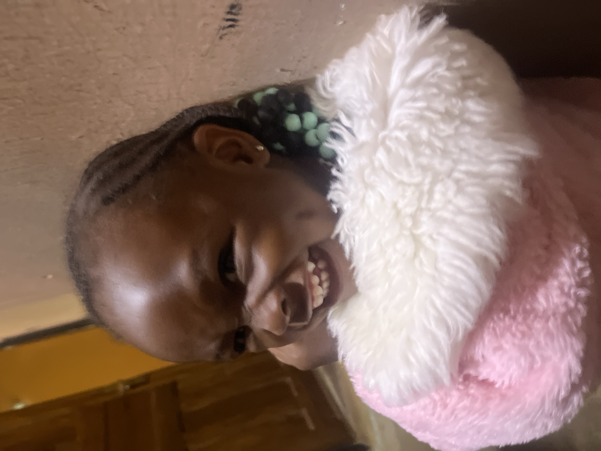 A close-up portrait shows a young Black girl smiling broadly, revealing her upper and lower teeth. Her dark hair is styled in neat braids, with several light green beads interwoven into the braids visible on the left side of her head. She wears a soft, fluffy garment, which is white on the upper part visible around her face and transitions to pink on the right side of her body. A small, golden earring is visible in her left earlobe. The background is a light tan or beige-colored wall, possibly textured, indicating an indoor setting. Faint, illegible markings or text are discernible on the wall in the upper left corner. The girl's joyful expression suggests a happy and playful moment. This scene takes place in Gura Top, Nigeria.