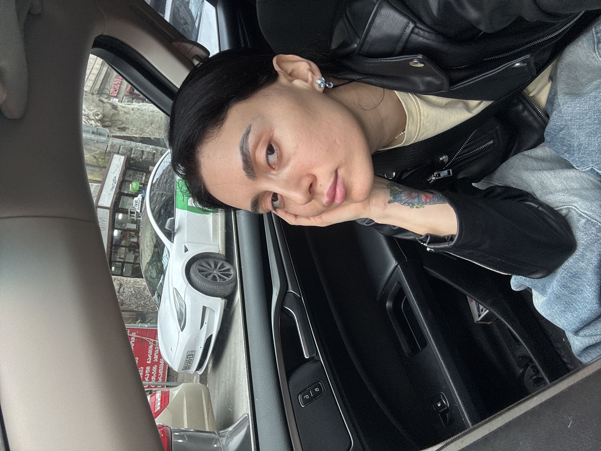 The image captures a female seated in the front passenger seat of a vehicle, her right hand supporting her chin as she looks towards the camera. She has dark hair, is wearing a black leather jacket over a light-colored shirt, and blue jeans. A multicolored tattoo is visible on her left forearm, and she wears blue and silver stud earrings. The interior of the car shows a black door panel with a silver handle, window lock controls, and a speaker grille. A small white and red box is present in the door's storage compartment. Through the car window, an urban street scene is visible. A white Tesla Model 3 taxi, bearing a Georgian license plate "GG-545-EB" and a green and white "TAXI" sign on its roof, is parked immediately outside. Parts of other vehicles are also present. The background features older stone buildings, trees, and storefronts with signage predominantly in Georgian script, including a partially visible "SUPERMARKET" sign. The scene is located on a city street in Tbilisi, Georgia.