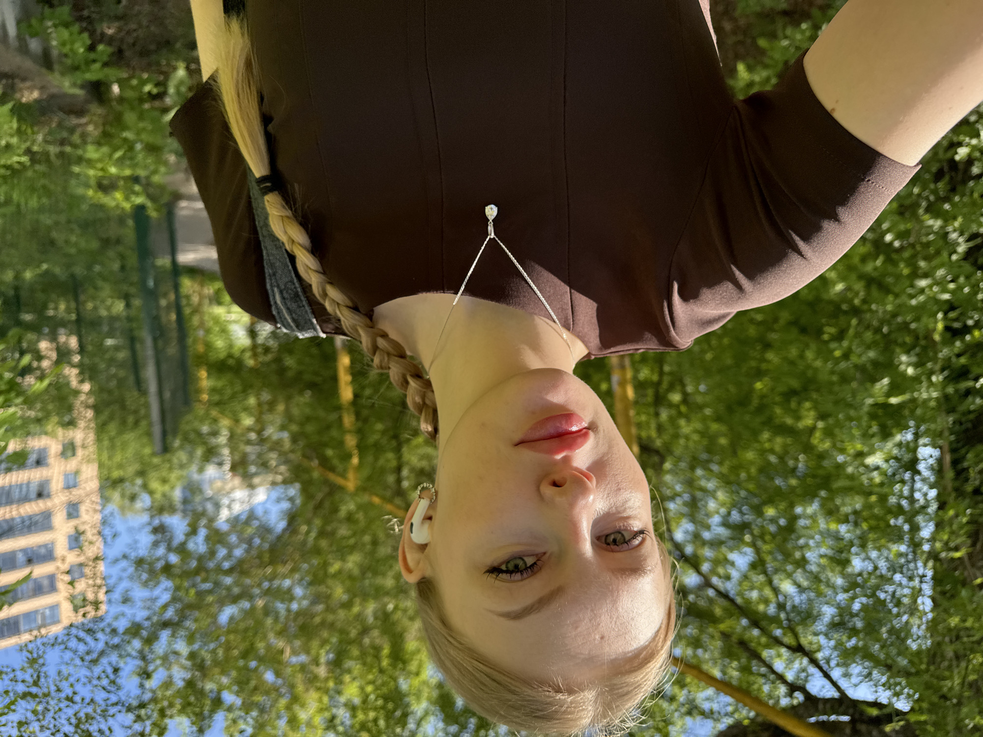 The image shows a woman with light skin, blonde hair in a braid, wearing a brown top and a silver necklace with a small pendant. She is also wearing white earbuds. The background includes green trees and the reflection of a building with multiple windows. Based on the reflections and foliage, the image was taken outdoors, possibly in a park or garden. The city location is Perm, Russia.