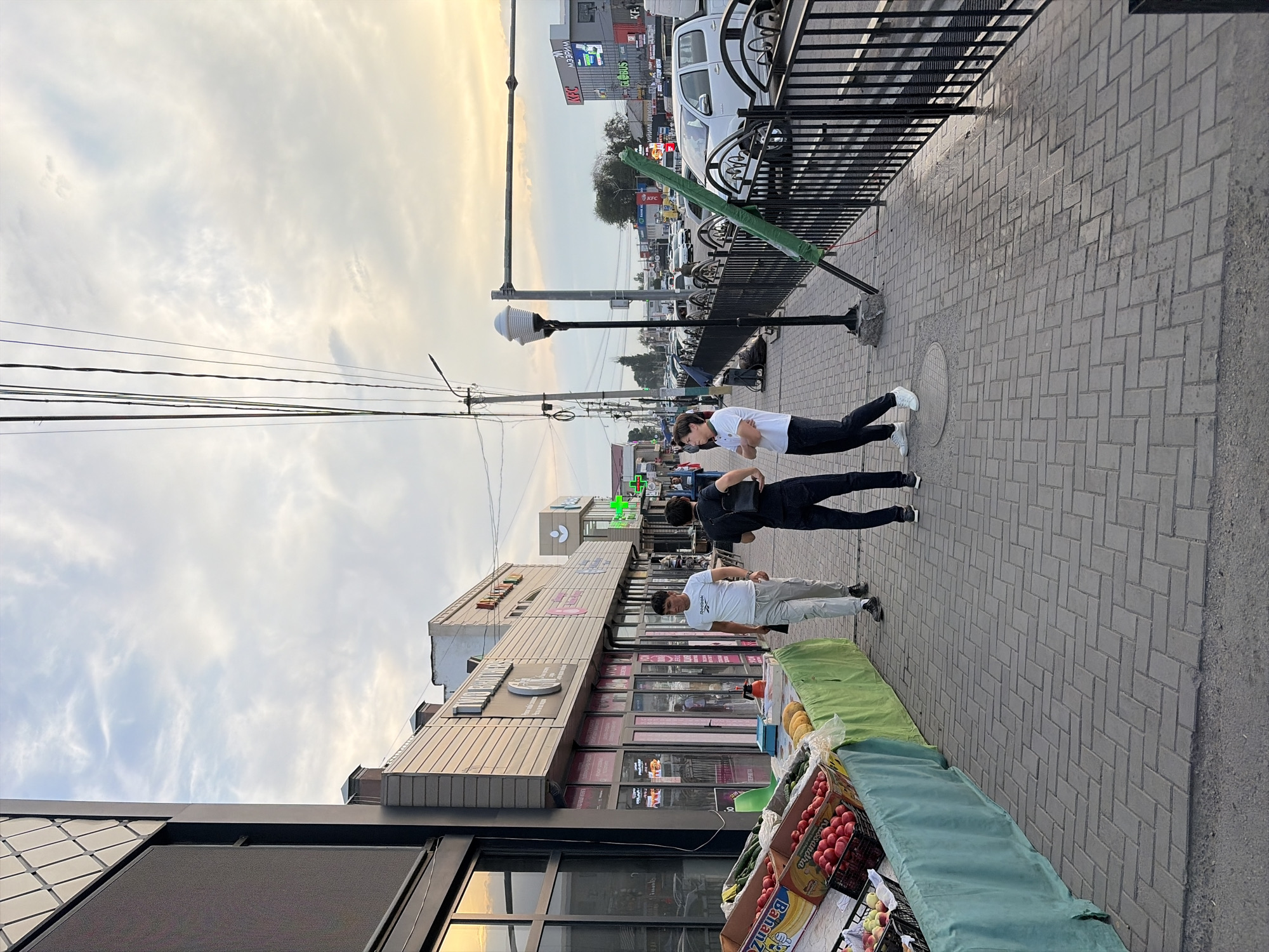 The image depicts a street scene in Kant, Kyrgyzstan, likely in the late afternoon or early evening due to the warm, diffused light. The sky is overcast with wispy clouds. A pedestrian walkway paved with gray bricks runs along the right side of the frame, bordered by a black metal fence. Beyond the fence, a street with parked cars and buildings is visible.
On the walkway, three young men are present. Two are standing closer to the foreground, facing away from the camera and towards the street. One is wearing a white t-shirt and black pants, with his arms crossed. The other, behind him, wears a black t-shirt and black pants, with a black bag slung over his shoulder. Further down the walkway, another man in a white t-shirt and light-colored pants stands facing the camera, looking down.
In the foreground, a makeshift market stall displays various fruits and vegetables, including boxes of tomatoes and bananas. The stall is covered with green cloths. In the background, commercial buildings line the street. One building has signage that includes "WIZZAR" and "KFC". Another building has a sign with what appears to be Cyrillic lettering. Streetlights and overhead wires are also visible. The overall mood is casual, with people going about their day.