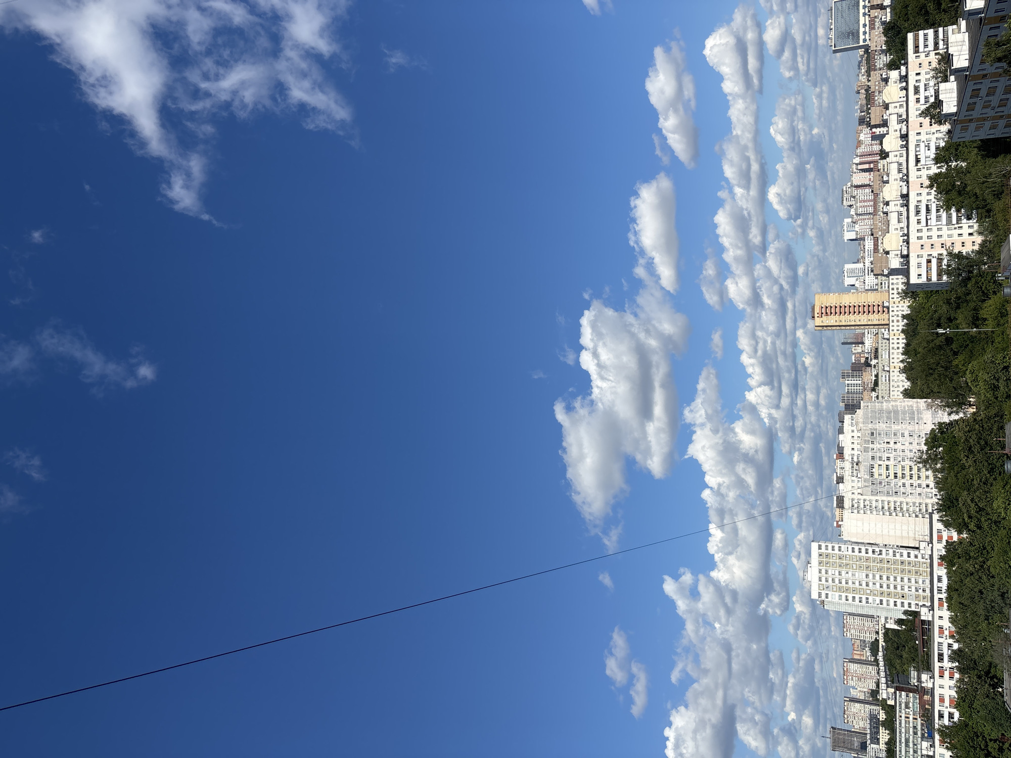 This is a photograph of a cityscape in Khimki, Russia, under a bright blue sky dotted with fluffy white clouds. The image captures a dense urban environment with numerous apartment buildings of varying heights and architectural styles. In the foreground, lush green trees create a natural border between the viewer and the buildings. A prominent thin black line, likely a cable, stretches diagonally across the upper left portion of the frame, from the top edge towards the center. The buildings are mostly residential, with many windows visible, suggesting a densely populated area. The overall impression is that of a typical day in a large Russian city, with clear weather and a clear view of the urban sprawl. There are no people or animals visible in the frame.
