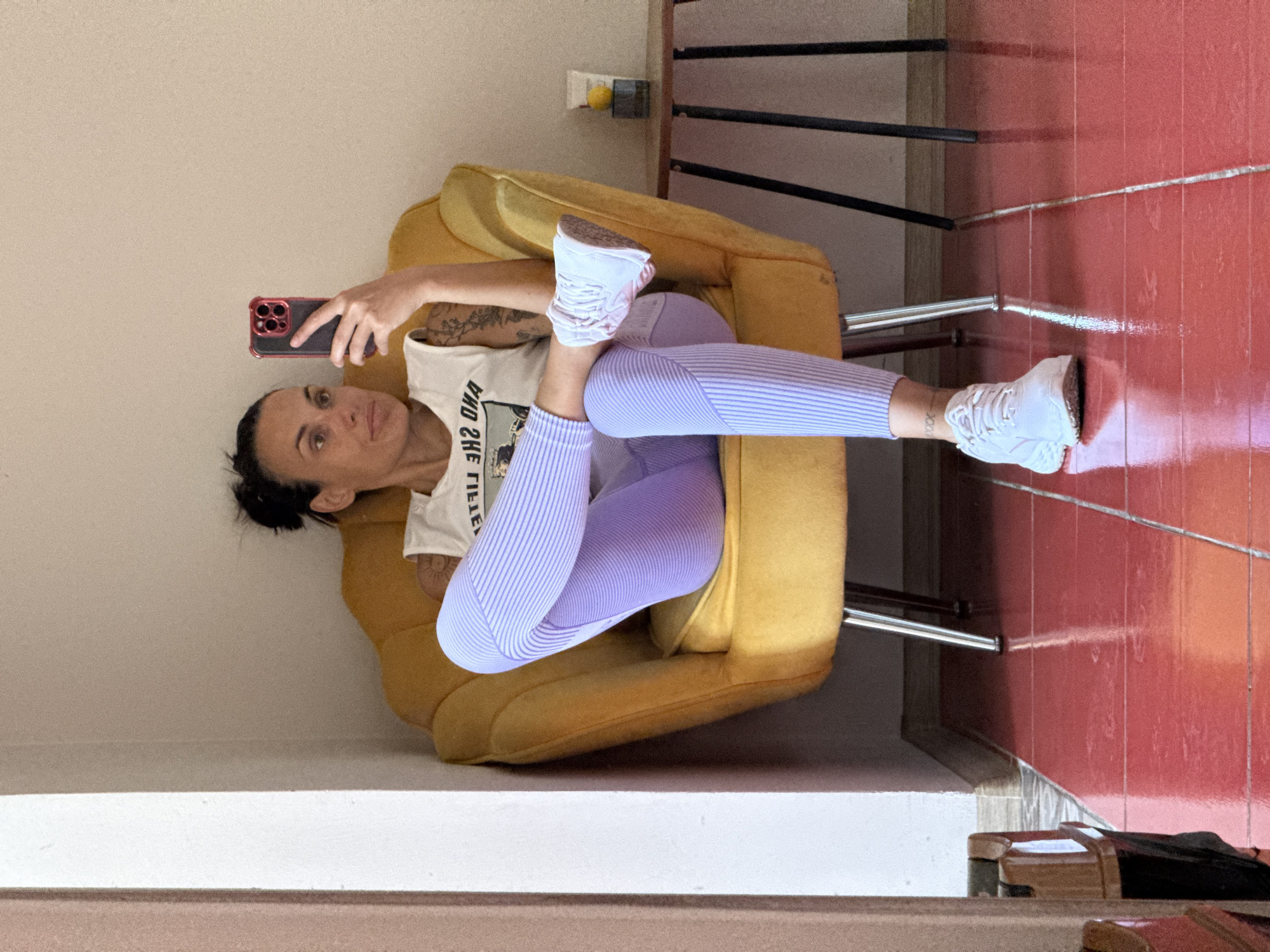 A woman is seated in a yellow armchair, taking a selfie with a smartphone. She is wearing a white tank top with a graphic print and light purple striped leggings. Her legs are crossed, and she is wearing white sneakers. The armchair has chrome legs and is positioned in front of a light-colored wall. To the right of the armchair, there is a small, dark wooden side table with a yellow object and a glass container on it. The floor is made of red tiles with a high gloss finish, reflecting the light and the furniture. The room appears to be indoors, possibly a living room or lounge area. There is a mirror to the left of the armchair, reflecting part of the scene. The overall impression is a casual, home environment. There is no information in the image to indicate the location is in the city of Pleven, Bulgaria.