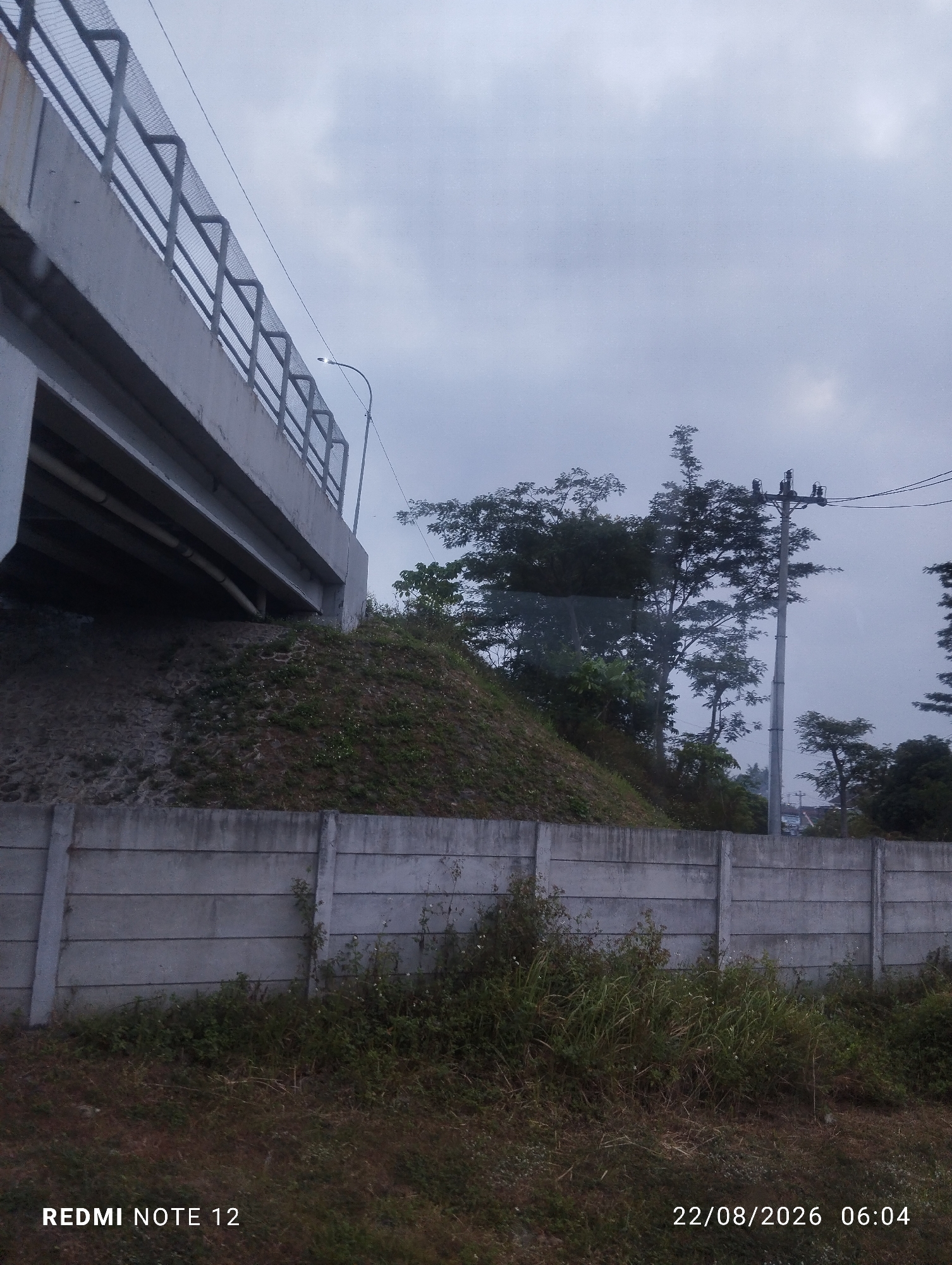 The image captures an overpass on a cloudy day. A concrete barrier topped with a metal fence runs along the edge of the overpass, which appears to be elevated and curves to the left. Below the overpass, a grassy embankment slopes down, partially obscured by a concrete retaining wall. Trees and vegetation are visible behind the wall, with a utility pole and power lines present in the background. The lower right corner of the image displays a timestamp reading "22/08/2026 06:04" and "REDMI NOTE 12". There are no people or animals visible in the scene.